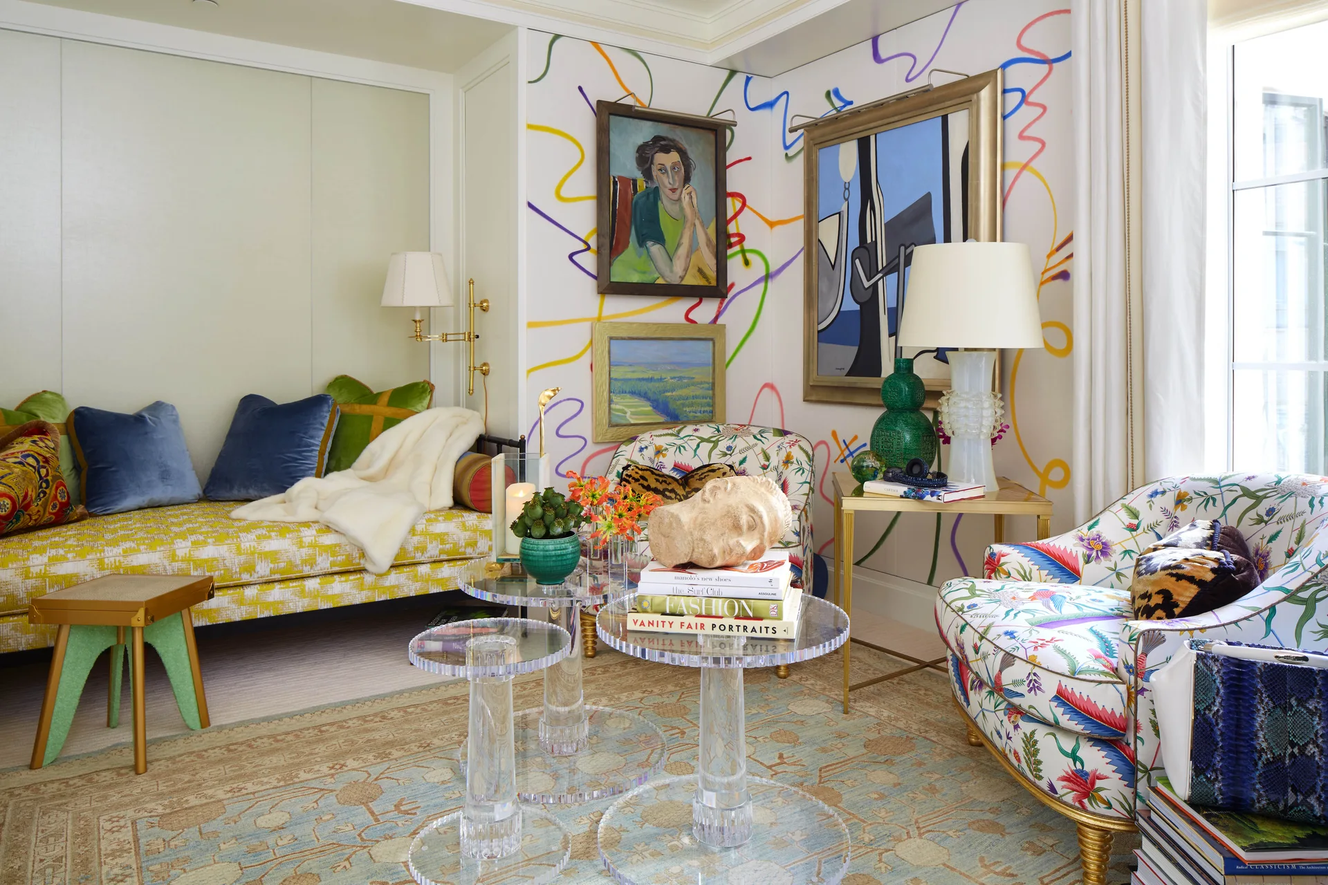 An eclectic living room with colorful squiggle-patterned wallpaper, a yellow daybed, floral armchairs, and clear acrylic nesting tables.