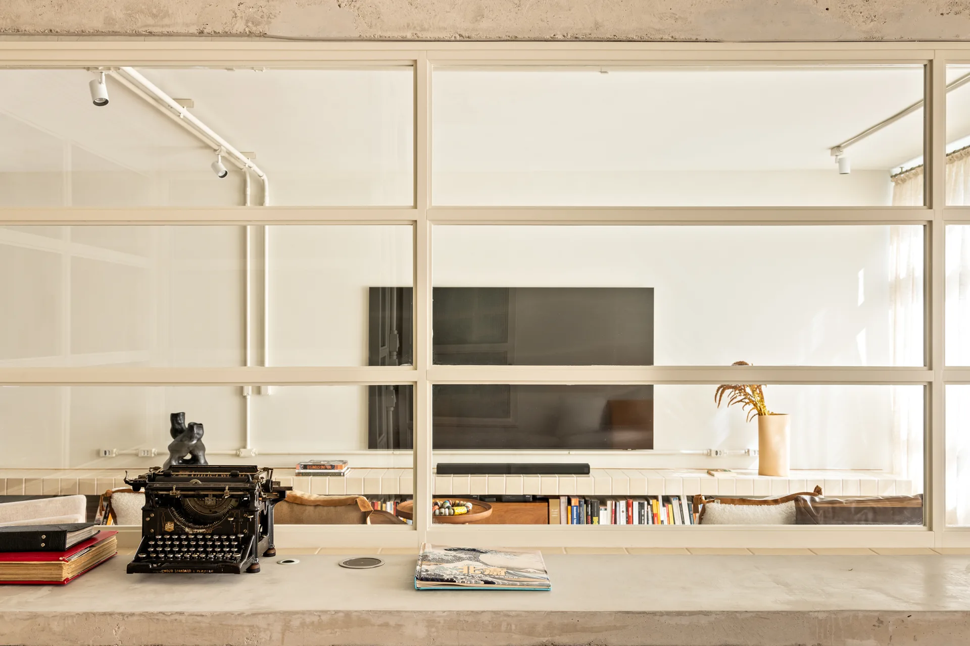 A view through a cream grid glass partition from a concrete desk with a vintage typewriter into a minimalist living room.