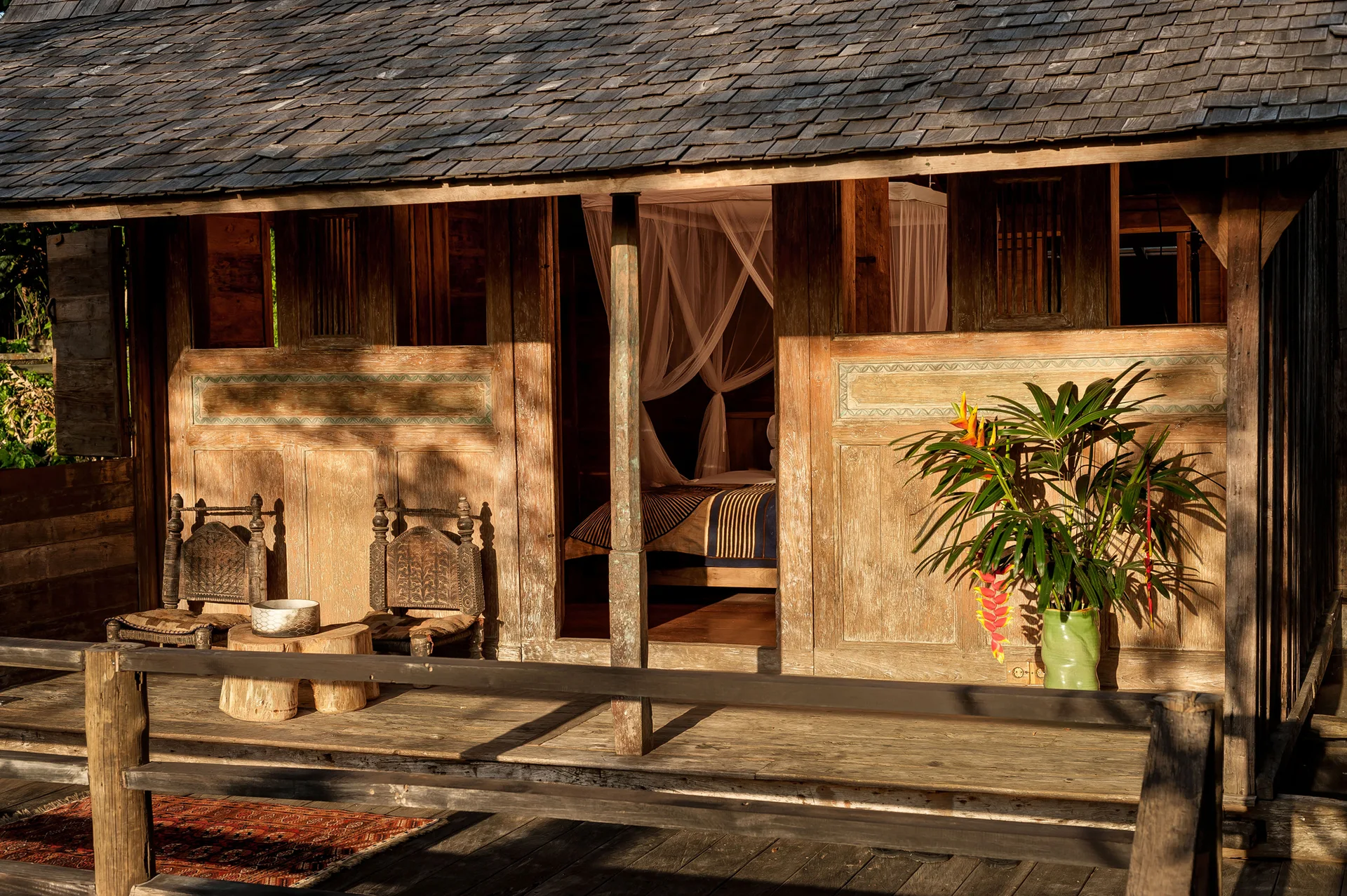 A rustic wooden porch features two carved chairs, a tree stump side table, and a wide doorway opening to a bedroom with a sheer canopy bed.