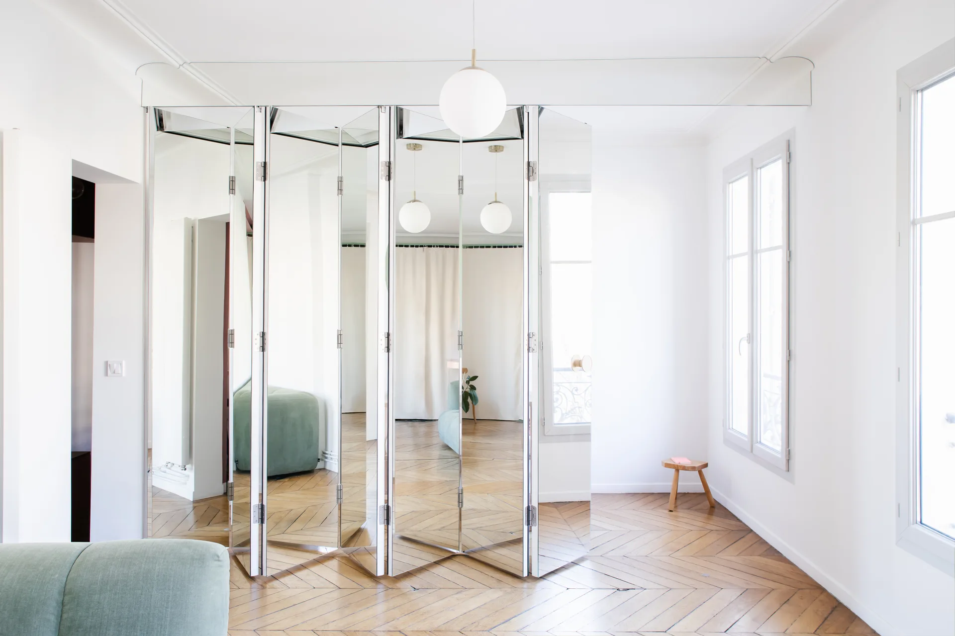 A bright white living room with herringbone parquet floors features a large mirrored folding screen reflecting the space.