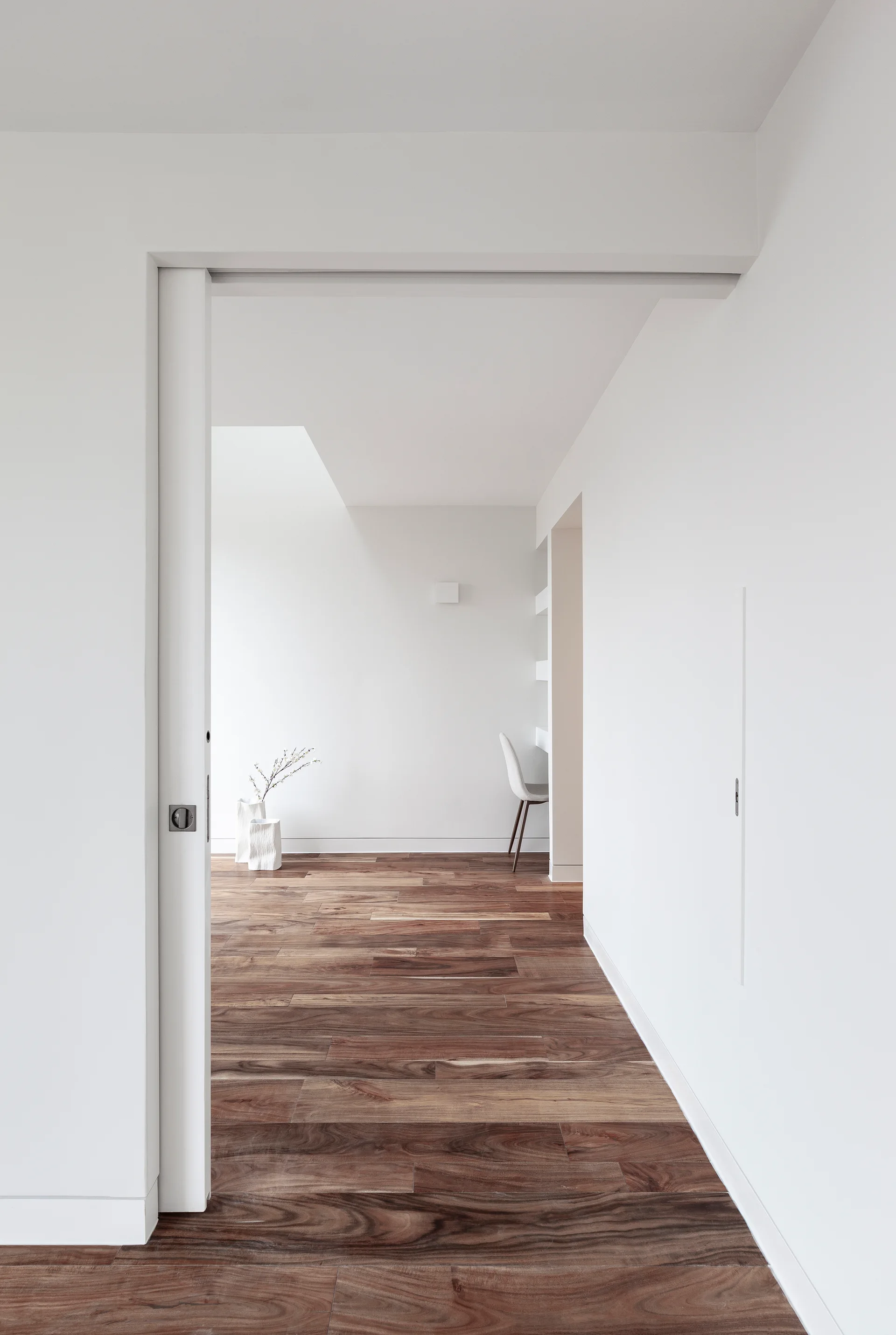 A minimalist white hallway features highly figured brown hardwood floors and an open sliding pocket door leading into a bright study.