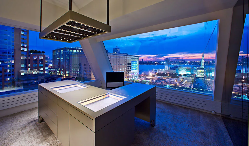 A modern grey office desk features two built-in glowing light panels under a suspended rectangular grid light fixture with a city view.