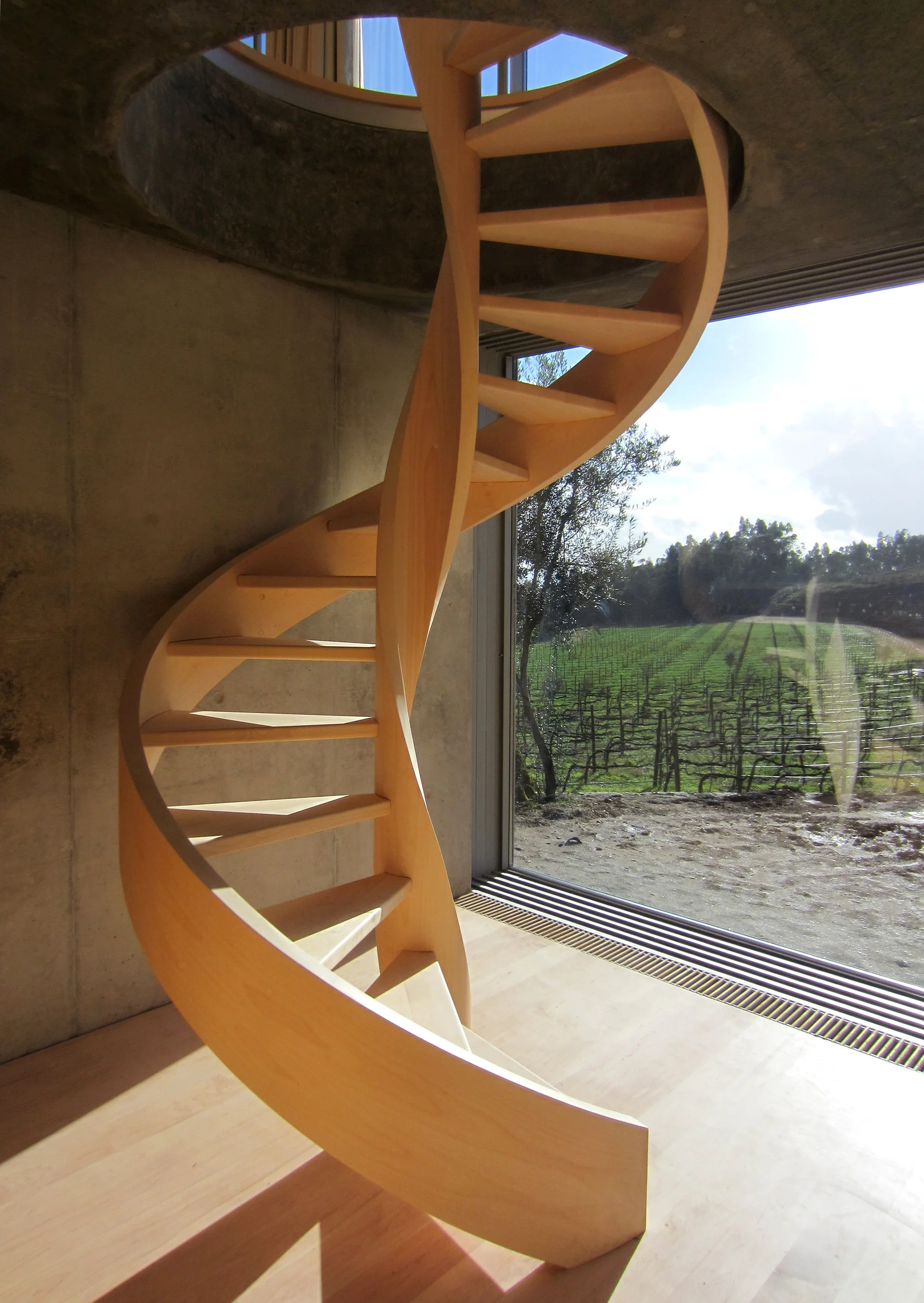 A light wood spiral staircase ascends through a circular ceiling cutout next to an exposed concrete wall and a large window.
