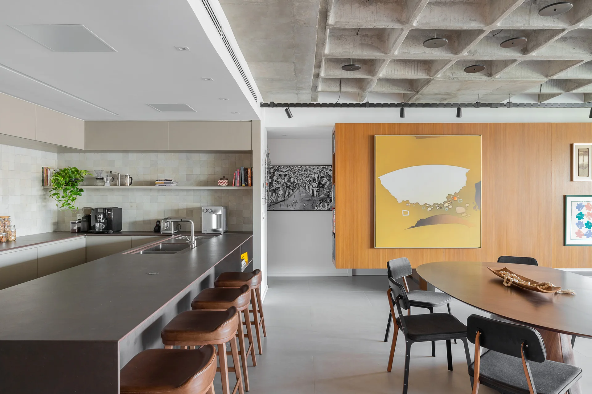 A modern open-plan kitchen and dining area featuring a concrete waffle ceiling, kitchen island with leather stools, and a wood-paneled wall.