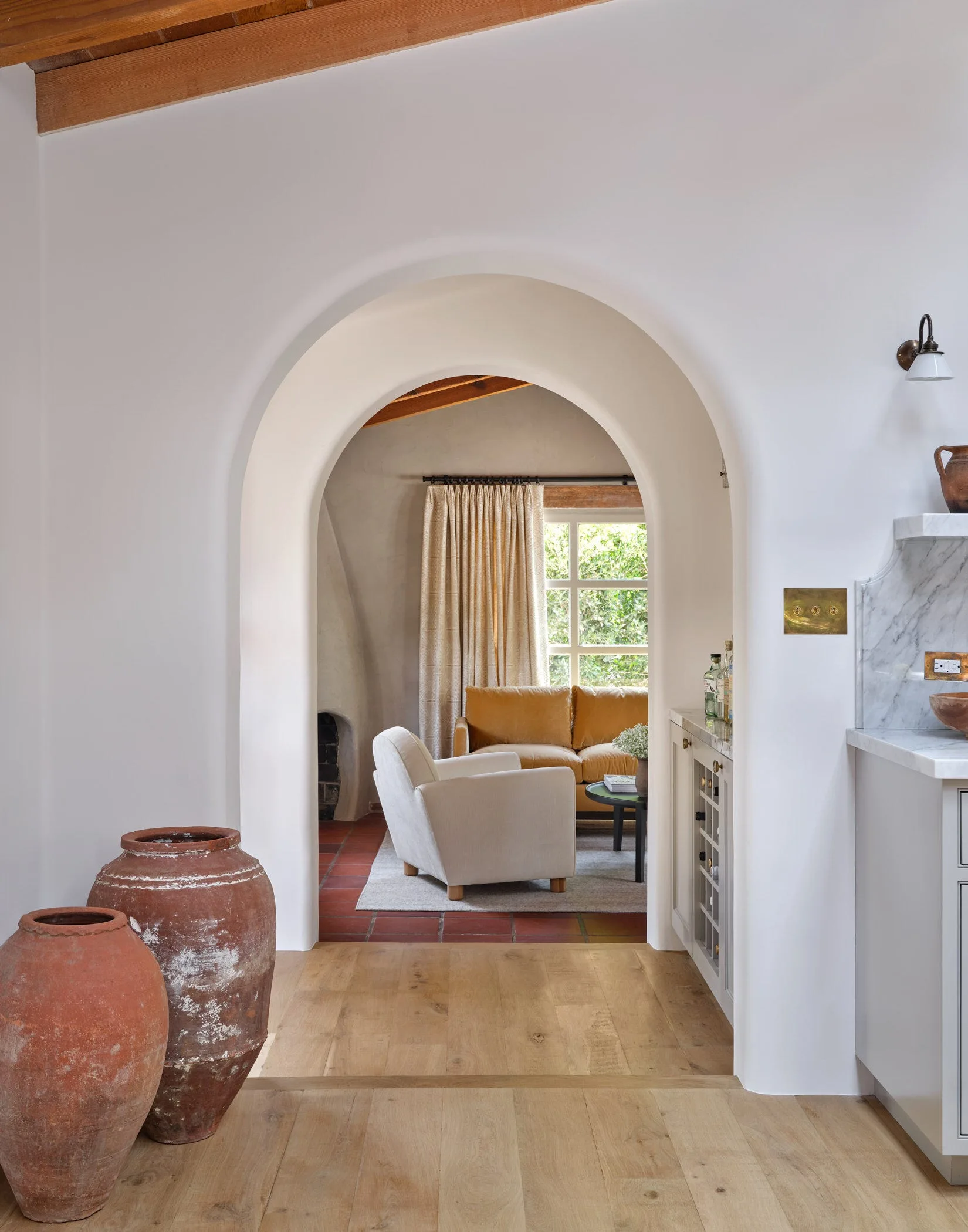 A white plastered archway frames a Spanish revival living room featuring a mustard yellow sofa, terracotta tile flooring, and rustic clay pots.