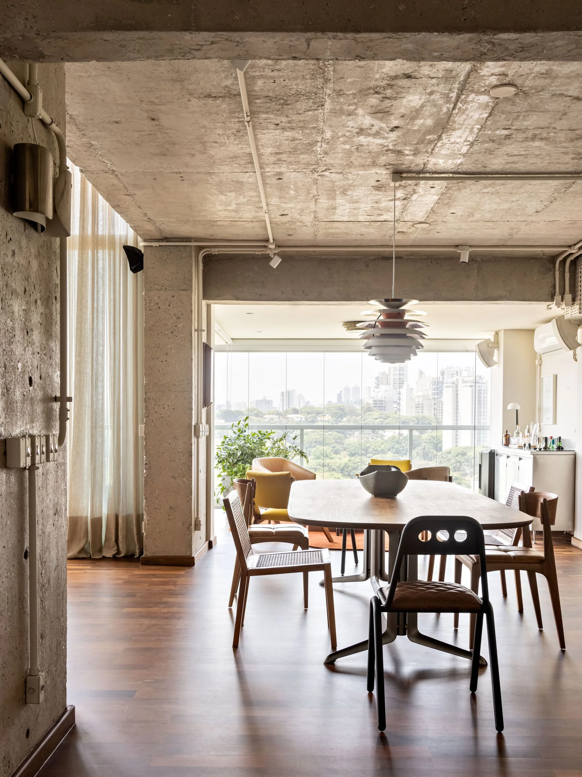 A modern dining room features a wooden table and mismatched chairs beneath an exposed concrete ceiling, with a city view through large windows.