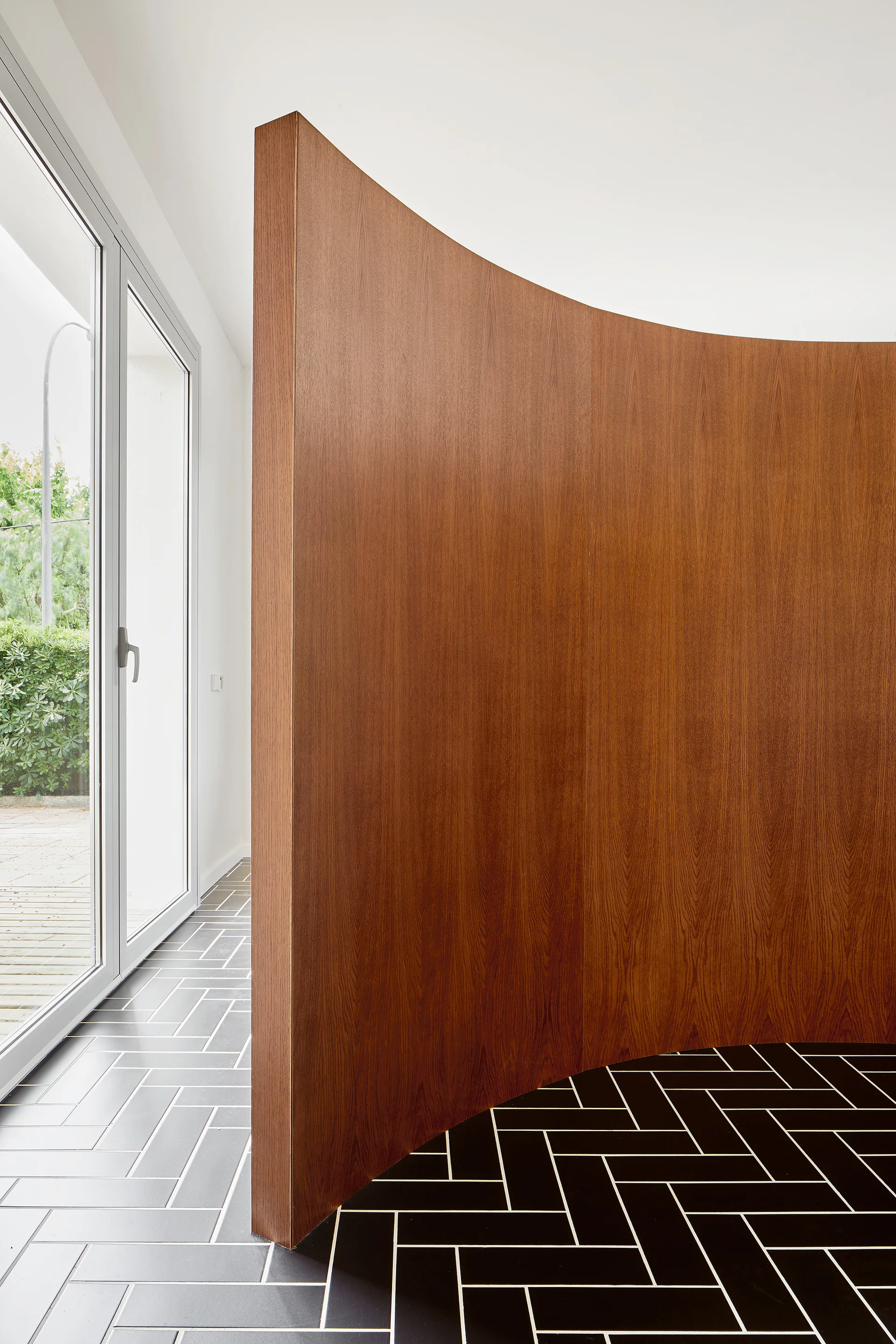A curved wood-veneered partition wall stands on a black herringbone tile floor next to a glass door.