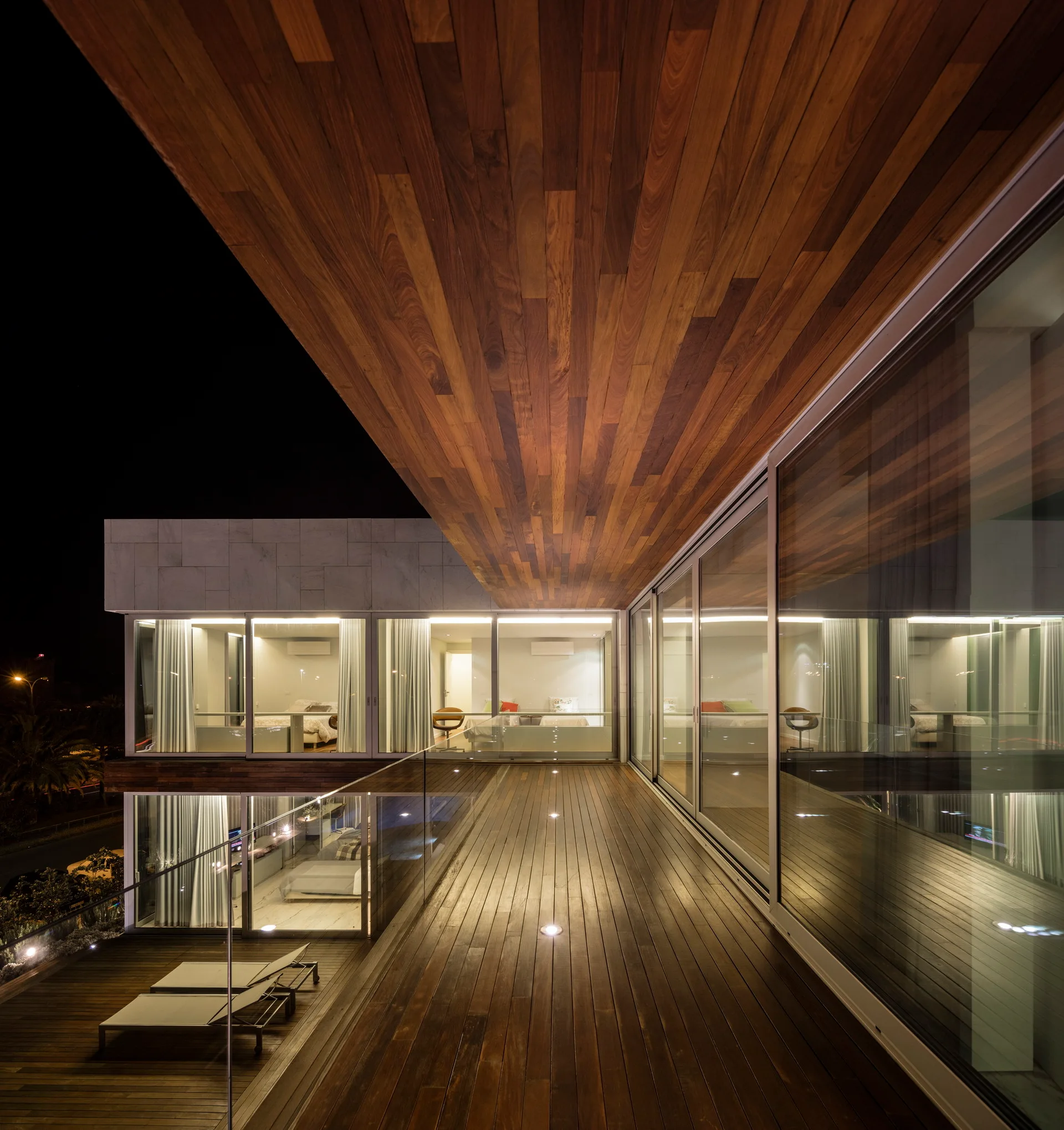 A modern residential exterior at night, showing a wooden balcony terrace with glass railings, warm floor uplighting, and large glass sliding doors.