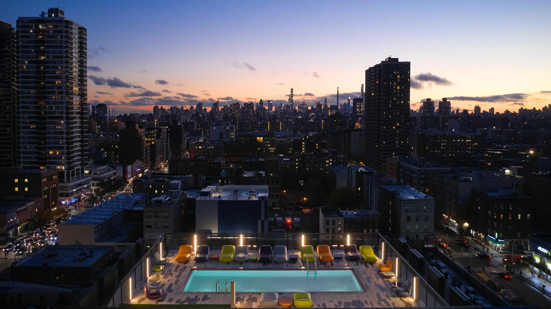 A rectangular rooftop swimming pool is illuminated at twilight, flanked by multicolored lounge chairs with a city skyline in the background.