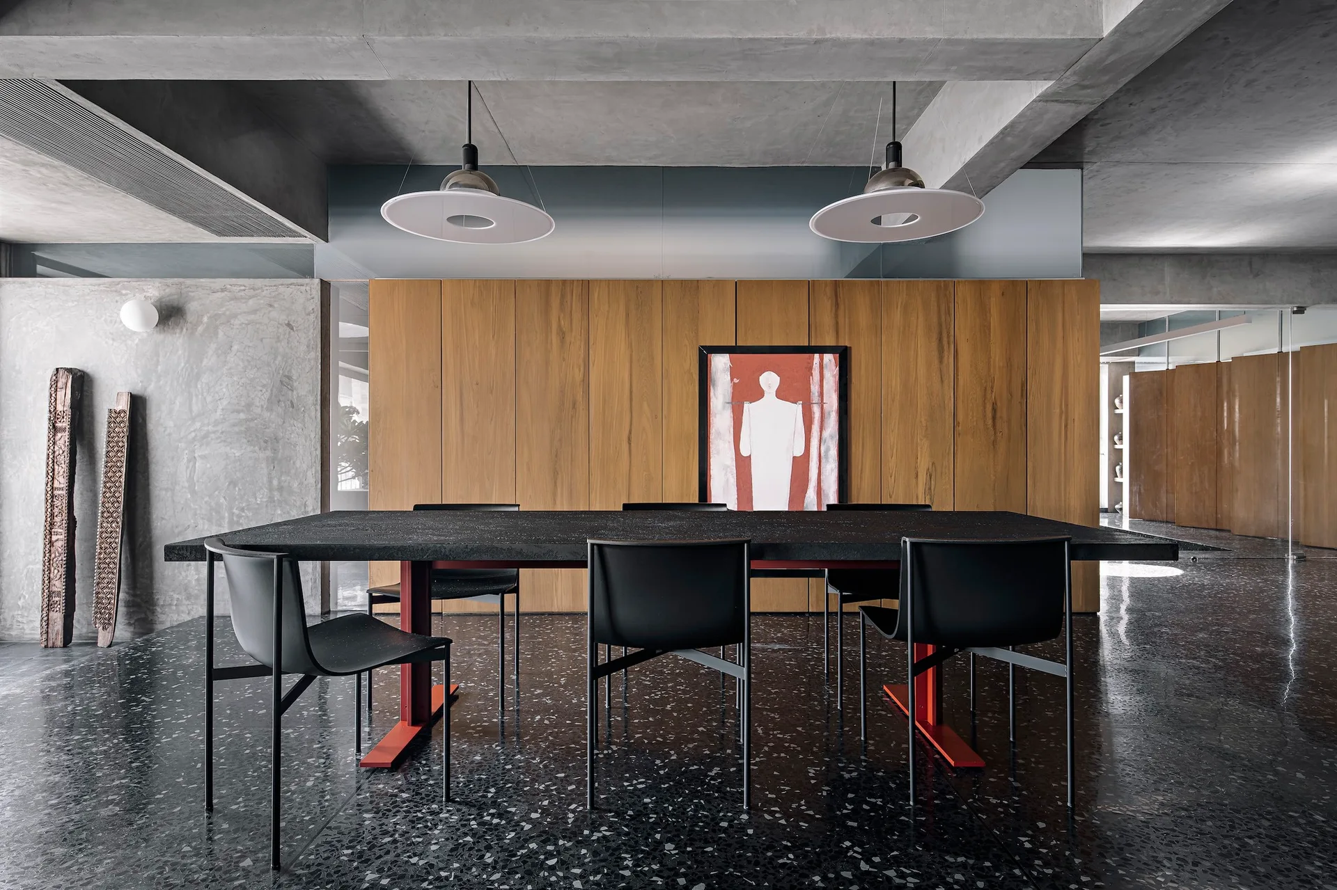 A modern dining room features a black table and chairs on a polished black terrazzo floor, set against warm wood paneling and a raw concrete ceiling.