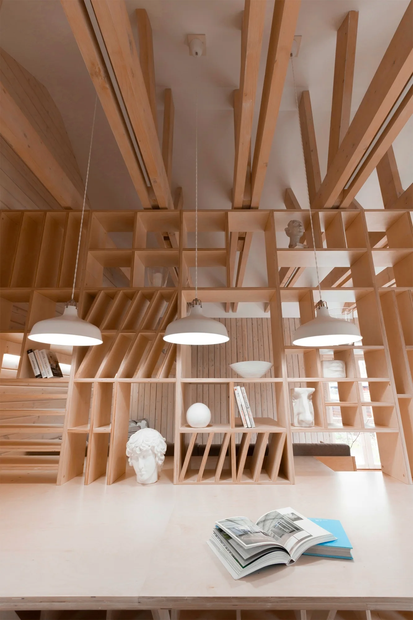 A custom light wood grid shelving unit serves as a room divider beneath exposed ceiling beams, illuminated by three white pendant lights.