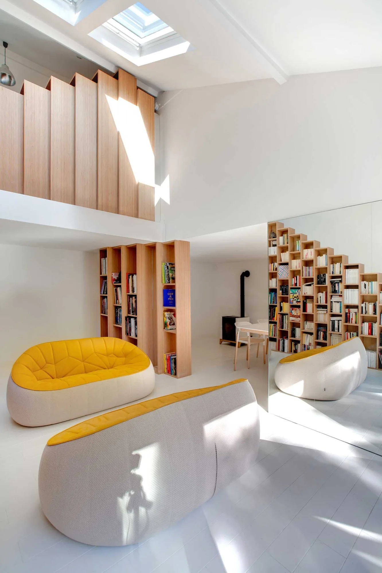 A bright minimalist living room with curved yellow and white sofas, a tiered light wood bookshelf, and a large wall mirror under a skylight.