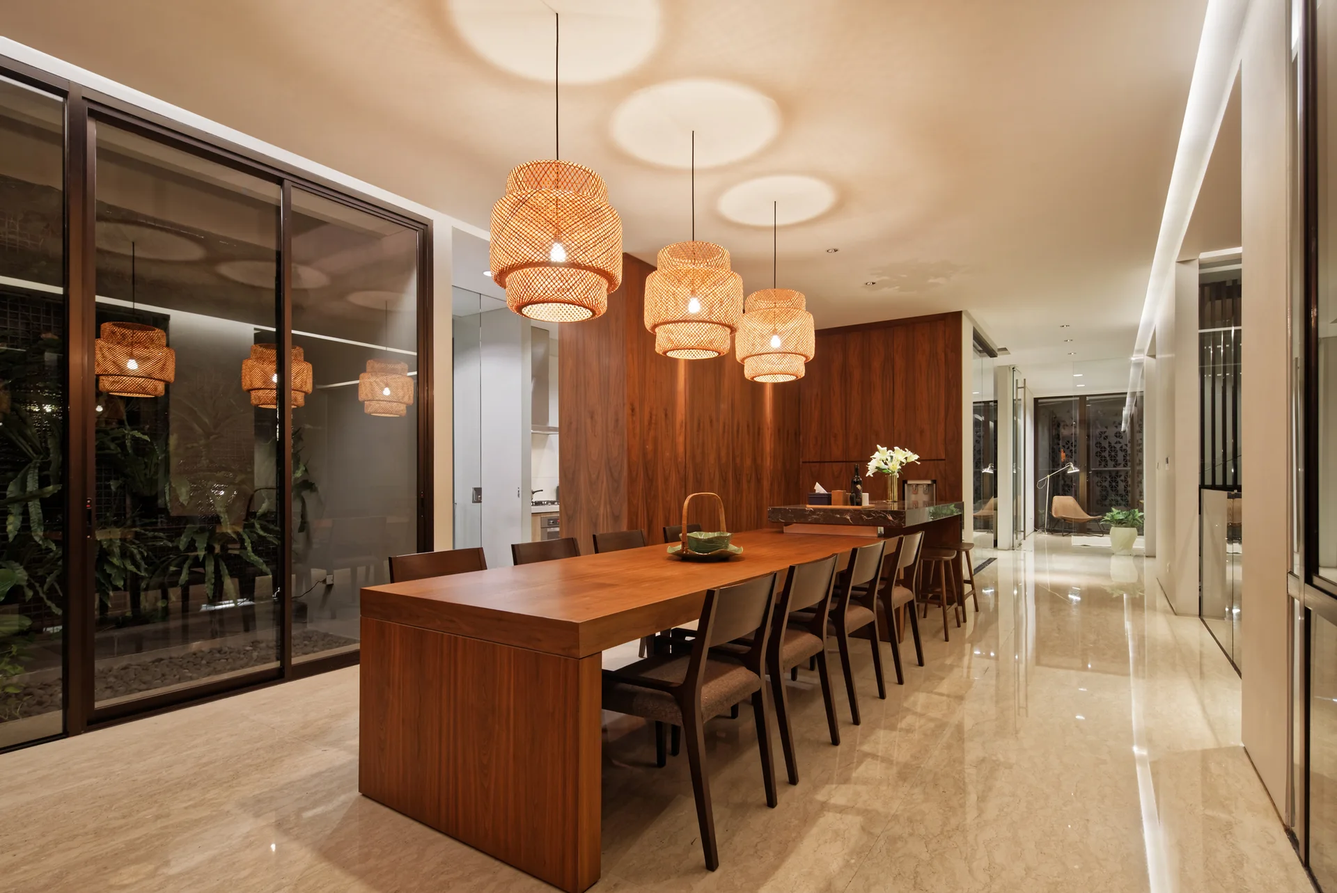 A long wooden dining table and chairs are arranged under three woven basket pendant lights in a room with wood-paneled walls and polished stone floors.