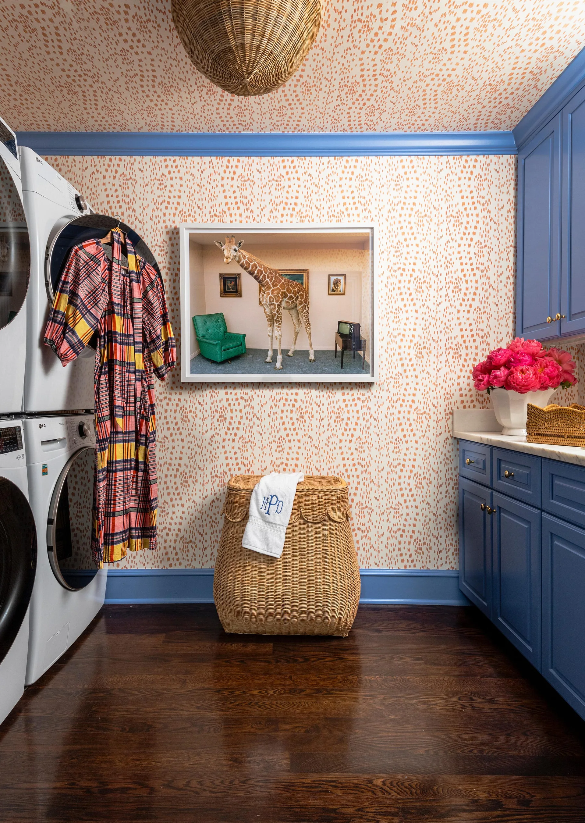 A laundry room features blue cabinetry, a stacked washer and dryer, orange-spotted wallpaper, a wicker hamper, and a framed giraffe art print.