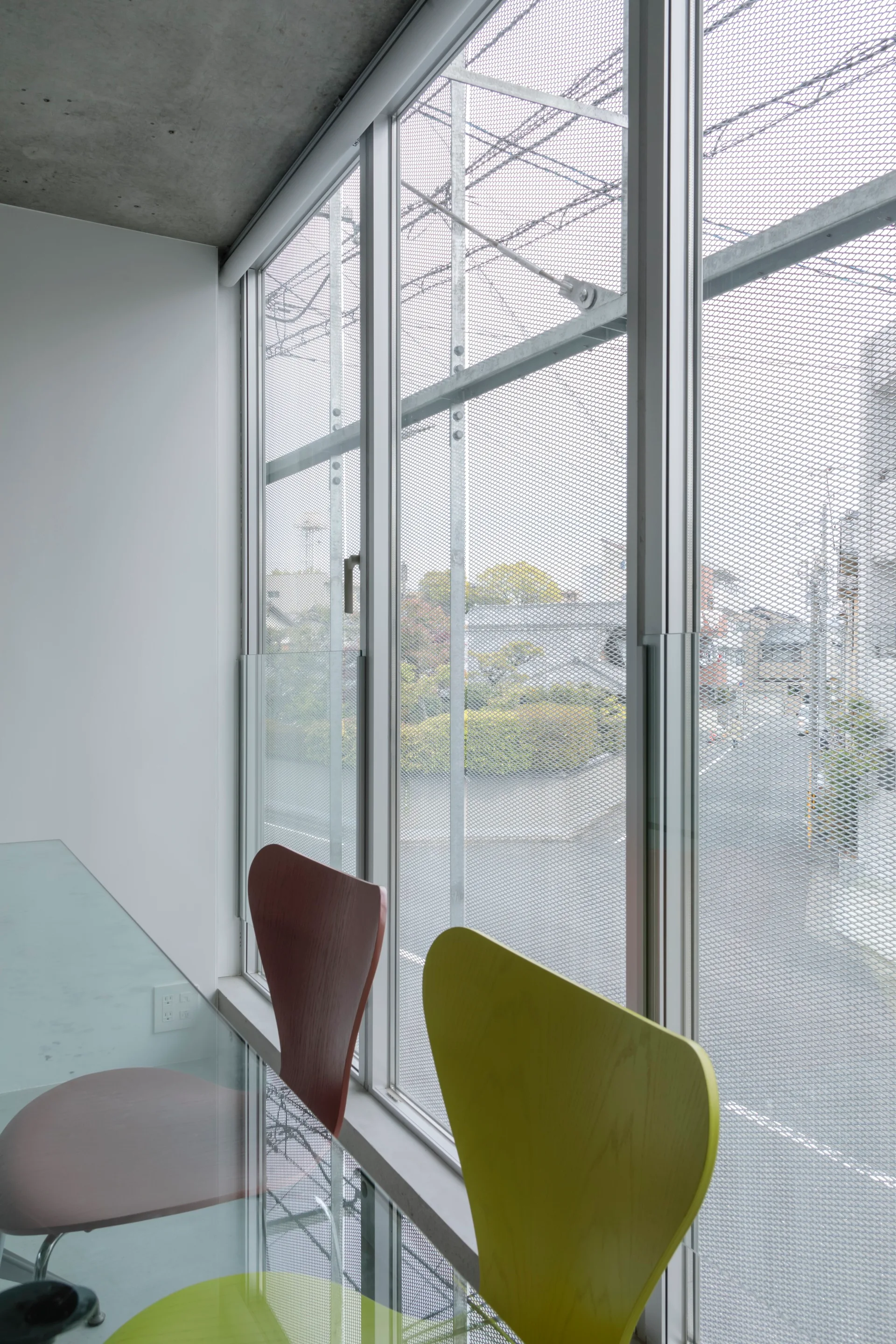 A minimalist office setting features a glass table with green and maroon wooden chairs next to a large window under an exposed concrete ceiling.