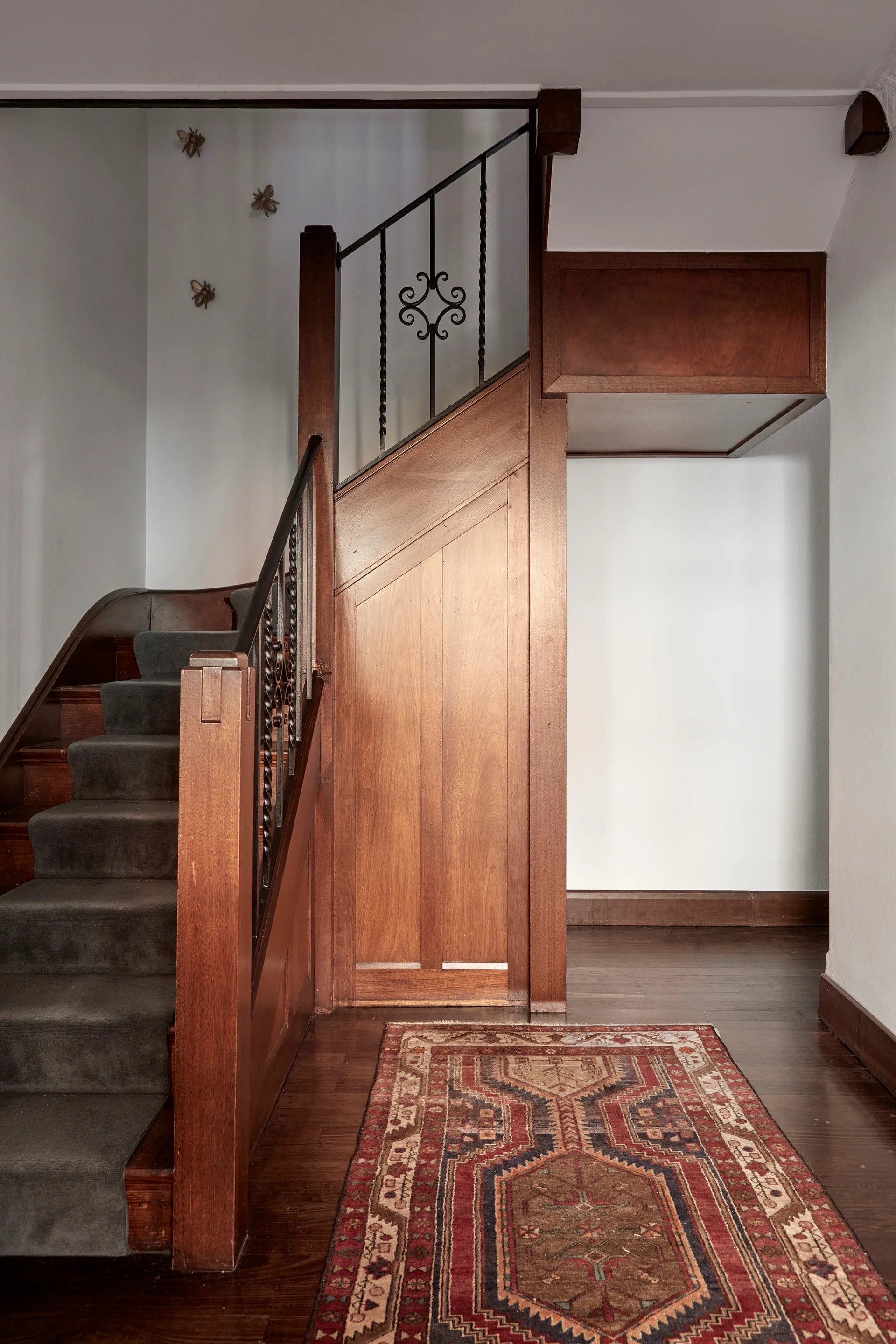 A stained wood staircase with a charcoal gray carpet runner, black wrought iron railing, and a patterned red rug on a polished hardwood floor.
