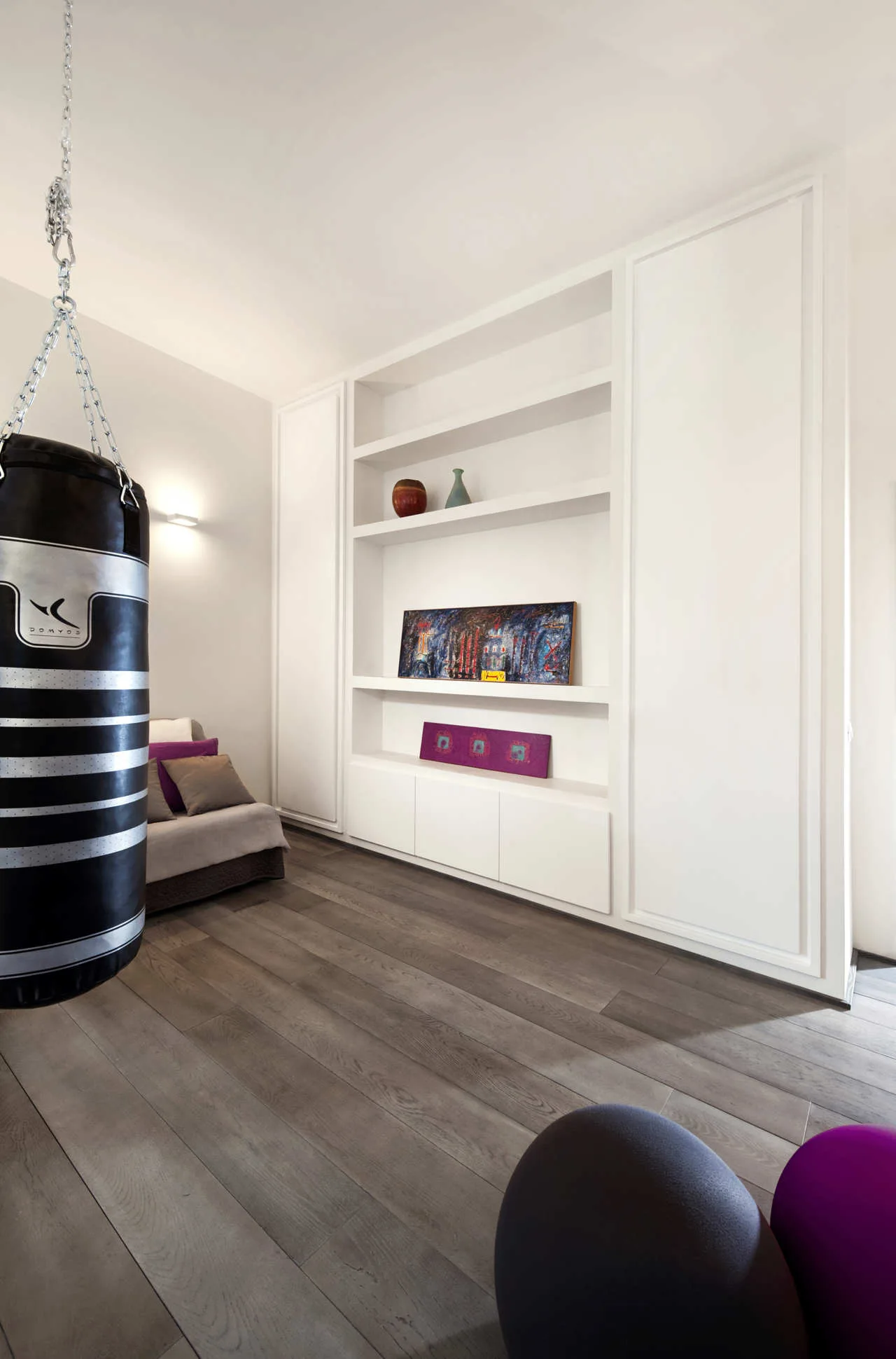 A black punching bag hangs from the ceiling in a modern home gym with white built-in cabinetry and dark wood floors.