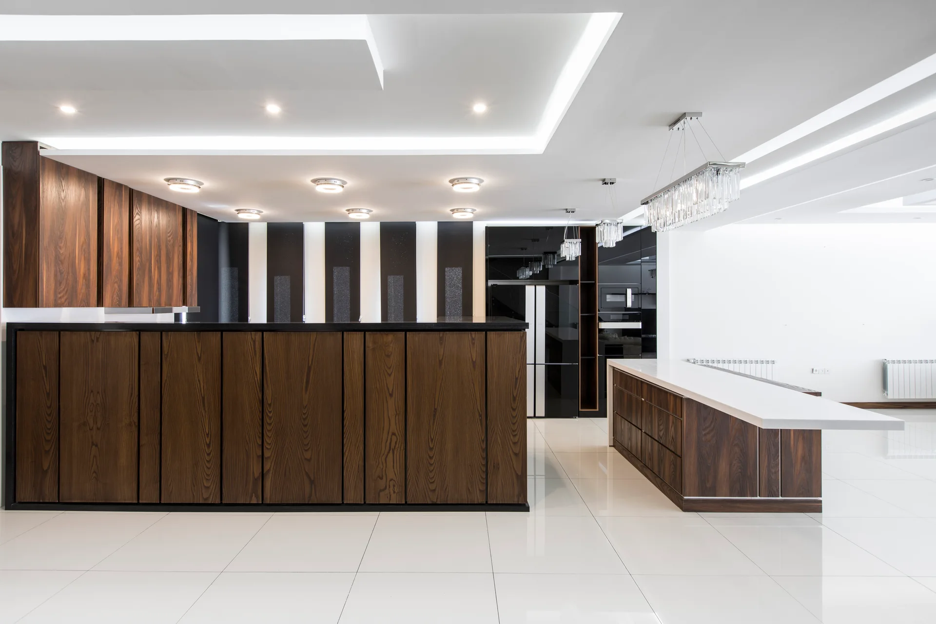 A spacious modern kitchen features dark wood grain cabinets, a matching island with a white countertop, and high-gloss white floor tiles.