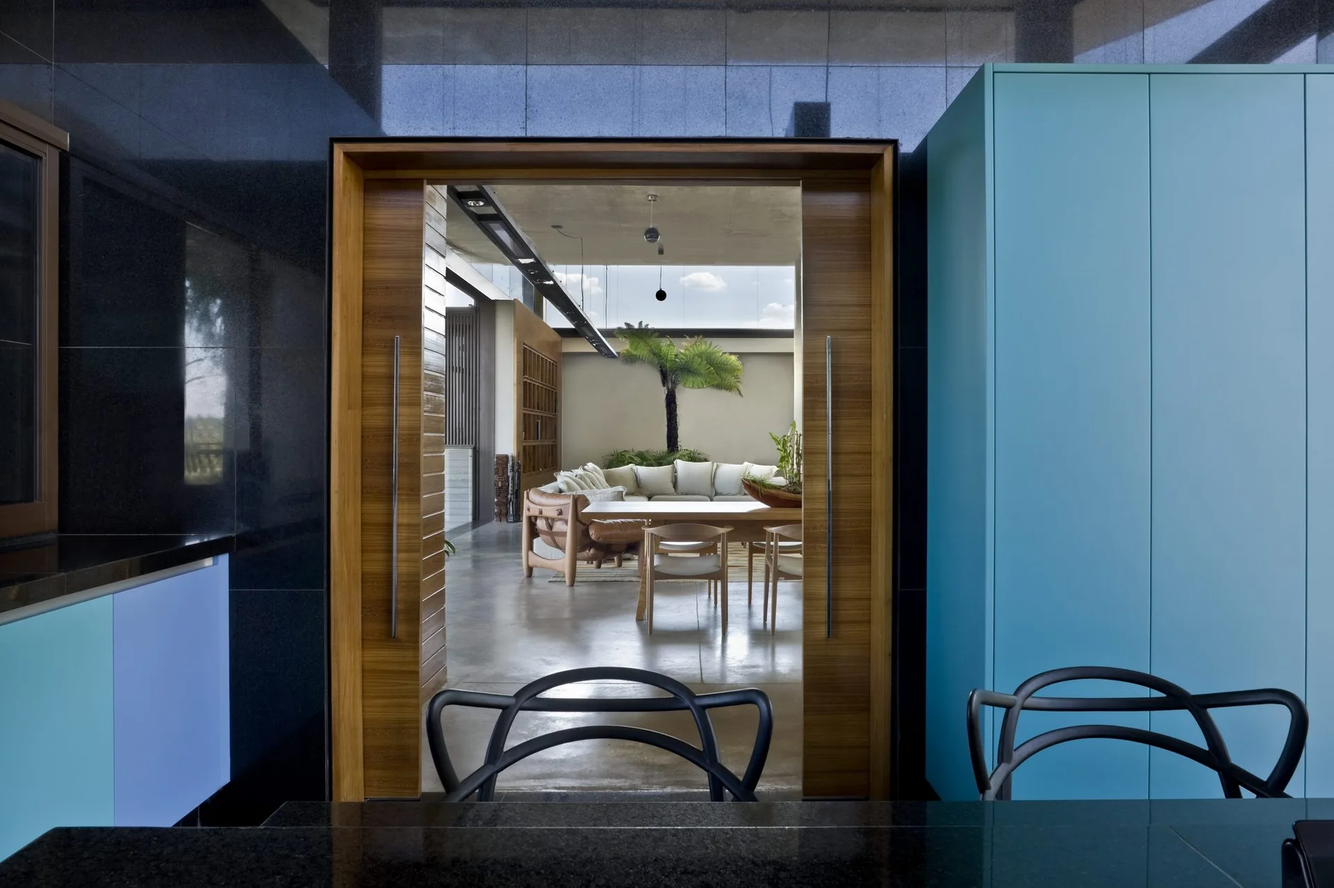 A wide wood-framed doorway offers a view from a dark foreground with blue cabinets into a bright, open-plan living room with a curved sofa.