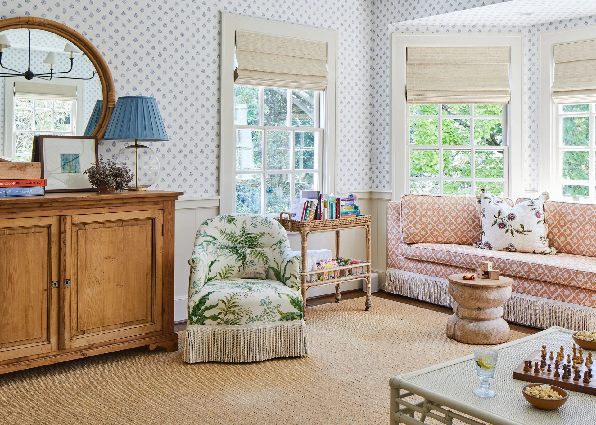 A traditional living room features blue patterned wallpaper above white wainscoting, an orange patterned sofa, and a floral armchair with bullion fringe.