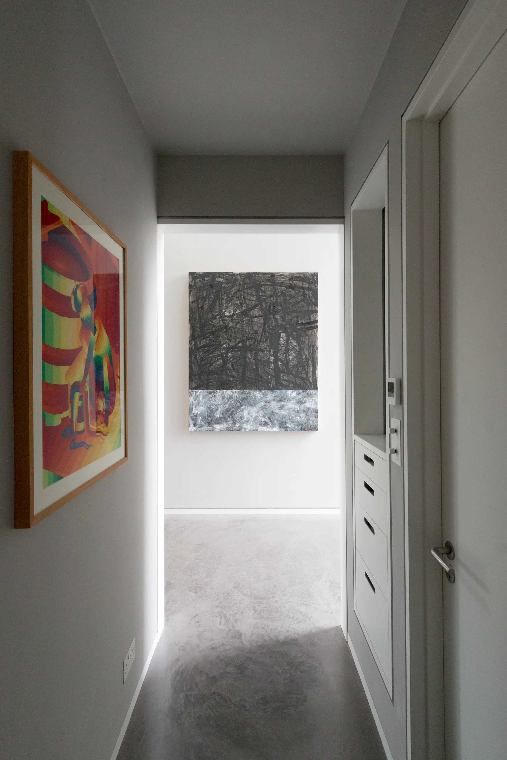 A narrow gray hallway with concrete floors features recessed white drawers, a colorful framed print, and a large abstract painting on the far wall.
