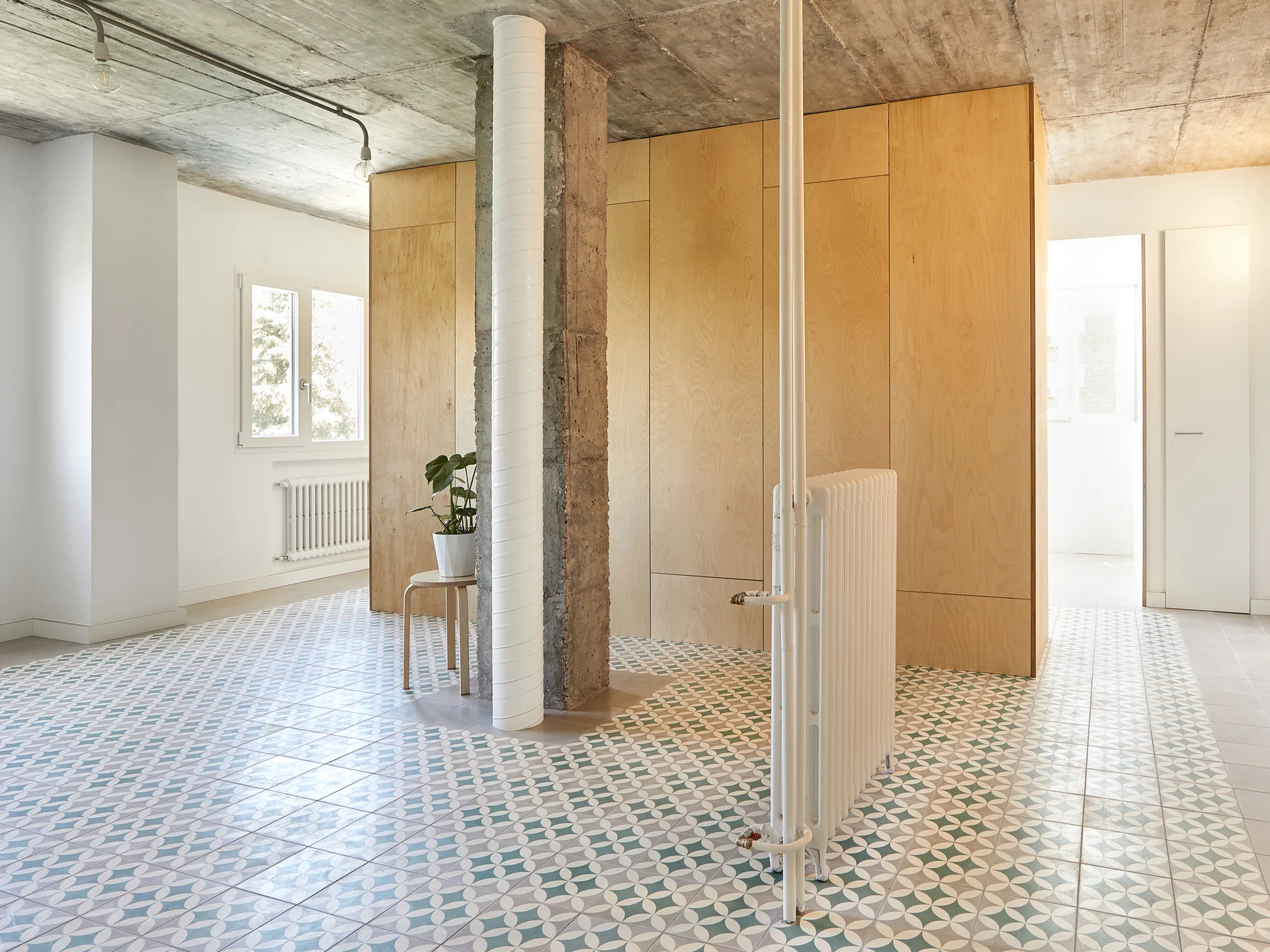 An open-concept room features green geometric patterned floor tiles, a plywood storage wall, an exposed concrete ceiling, and a central pillar.