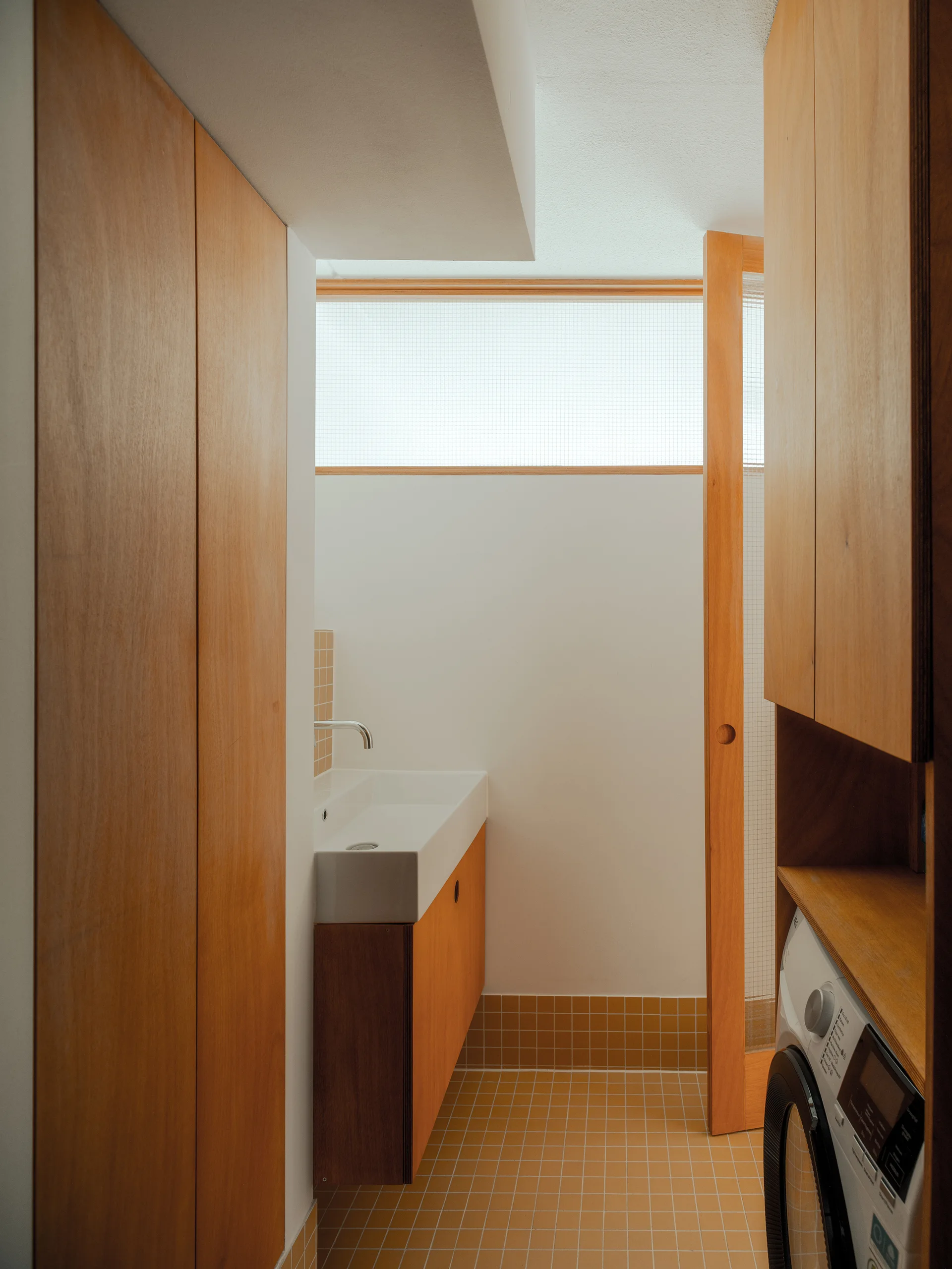 A minimalist utility room features warm wood cabinetry, a wall-mounted sink, a built-in washing machine, and ochre floor tiles.
