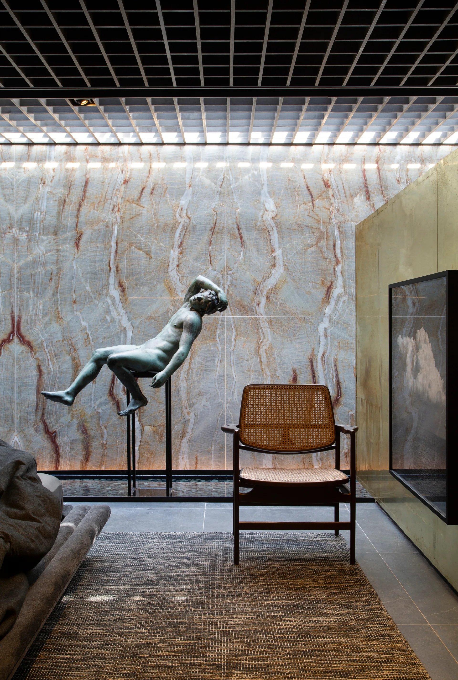 A modern living space featuring a backlit stone feature wall behind a bronze sculpture, a rattan accent chair, and a black grid ceiling.