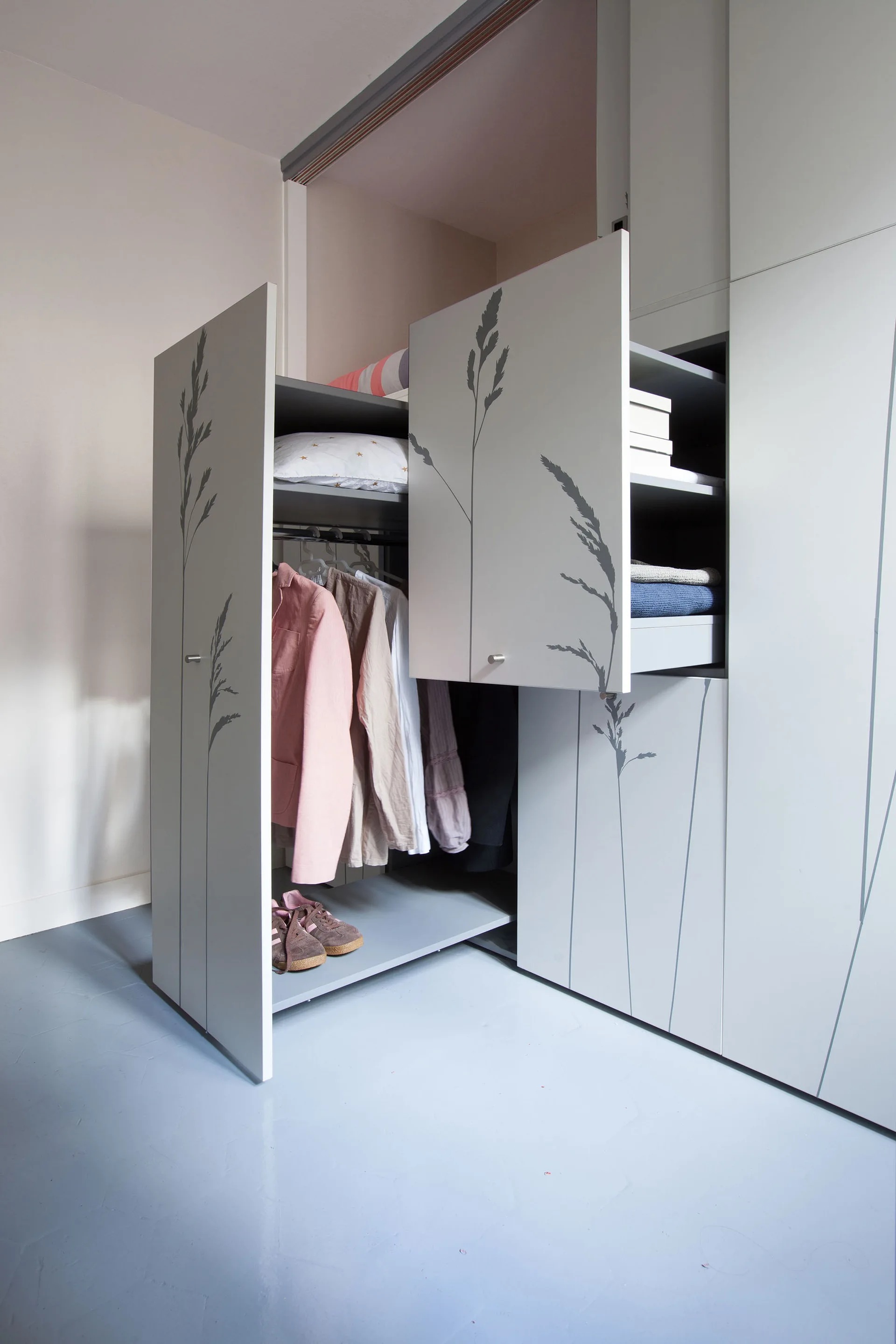 A modern grey wardrobe with open sliding doors featuring a painted grass pattern, revealing hanging shirts, folded clothes, and shoes inside.