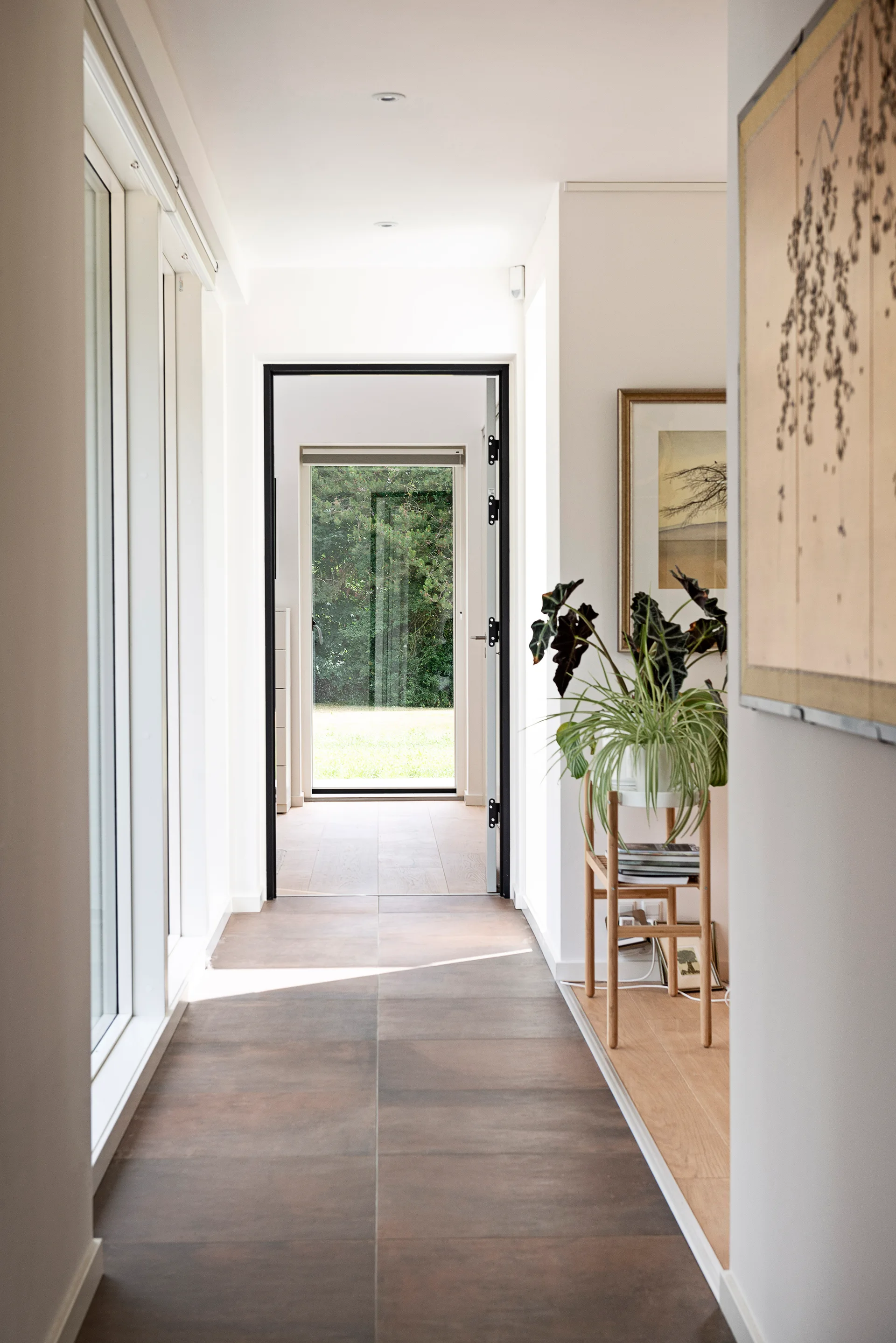 A long minimalist hallway features white walls, dark brown floor tiles, and a black-framed doorway looking out toward a green garden.