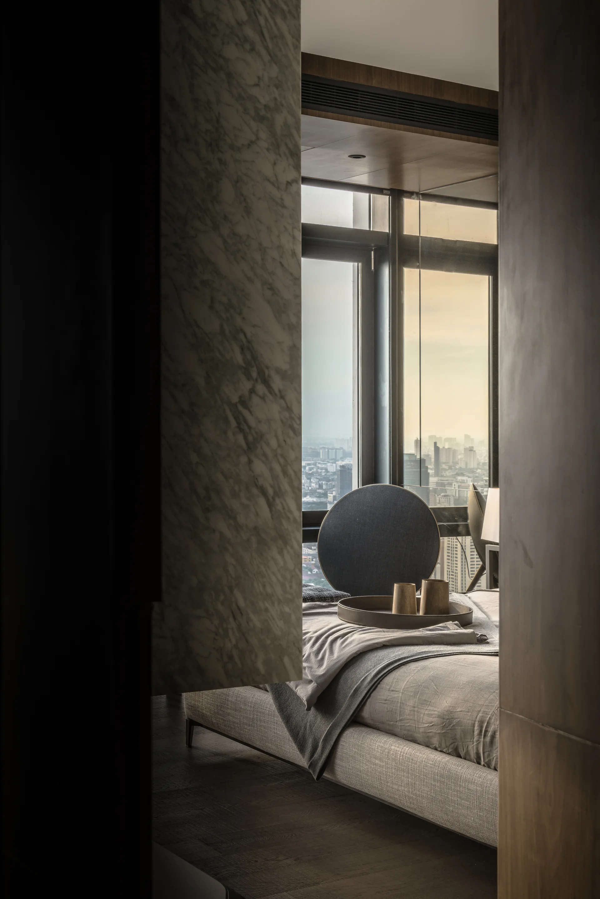 A doorway frames a modern bedroom with an upholstered bed, a marble wall panel, and a large window showing a city skyline.