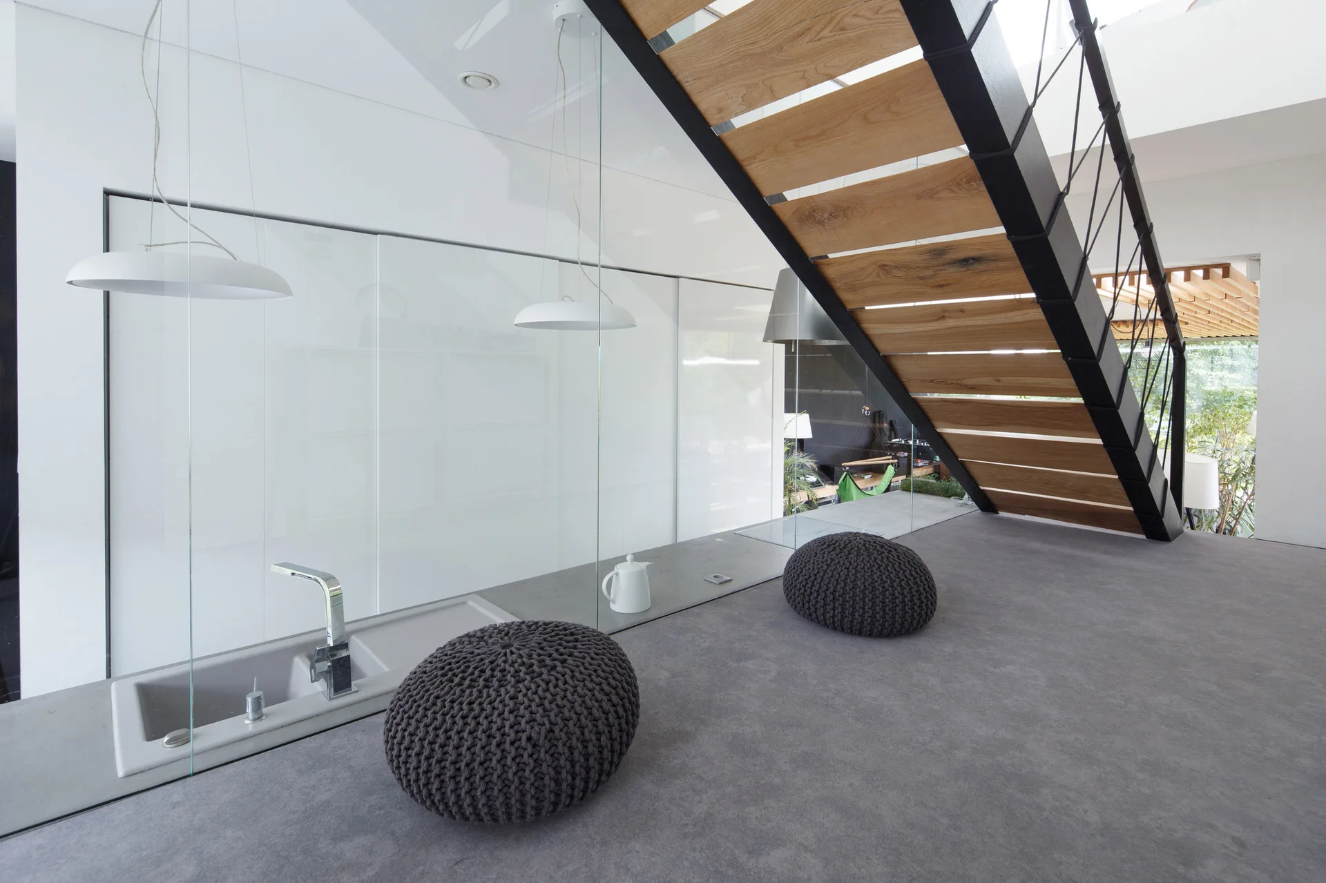 An open-concept modern interior features a floating wooden staircase, a grey built-in sink counter behind a glass partition, and two dark knitted poufs.