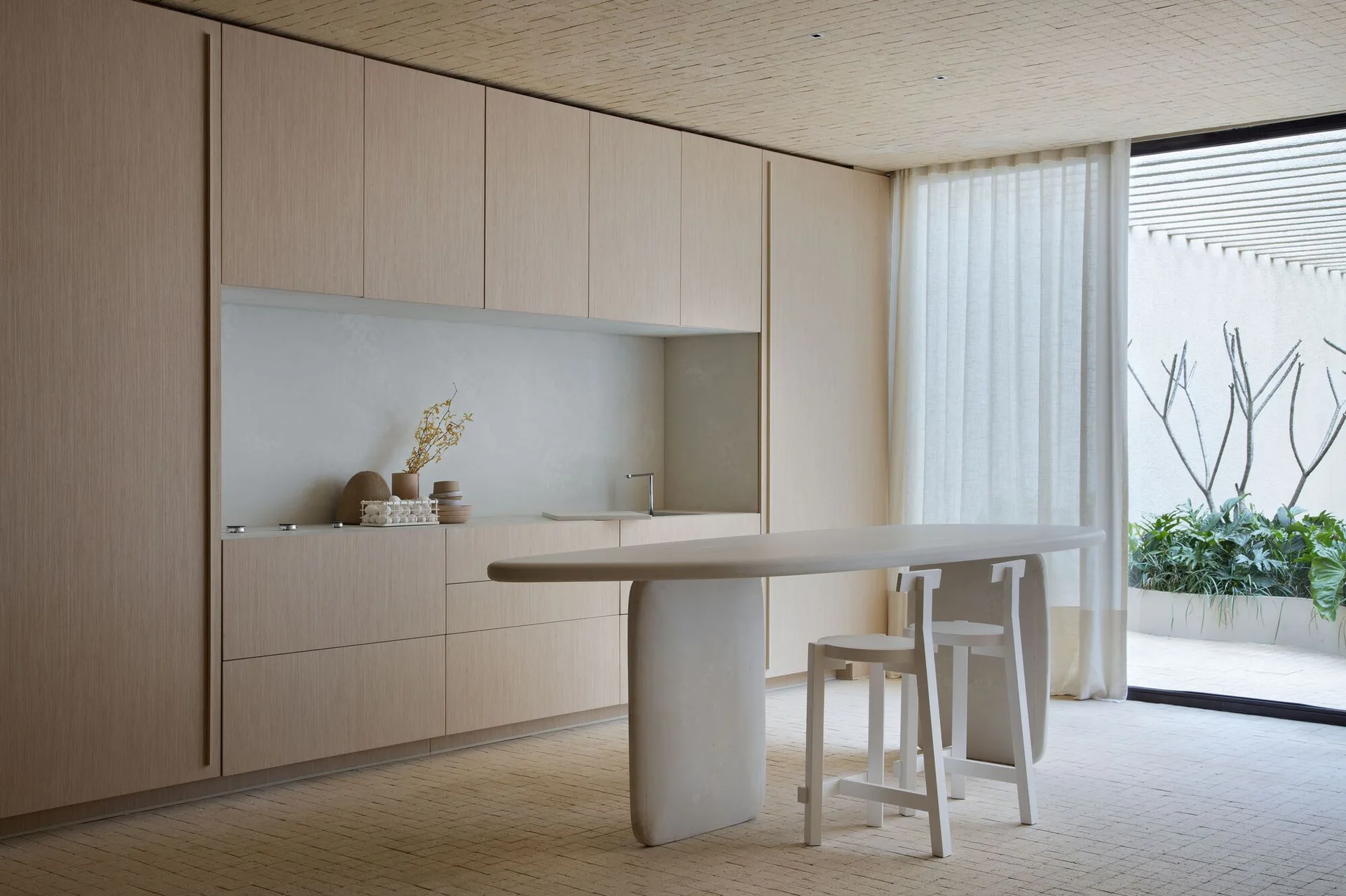 A minimalist kitchen features flat-panel light wood cabinetry, a sculptural beige island with two white stools, and matching tiled floor and ceiling.