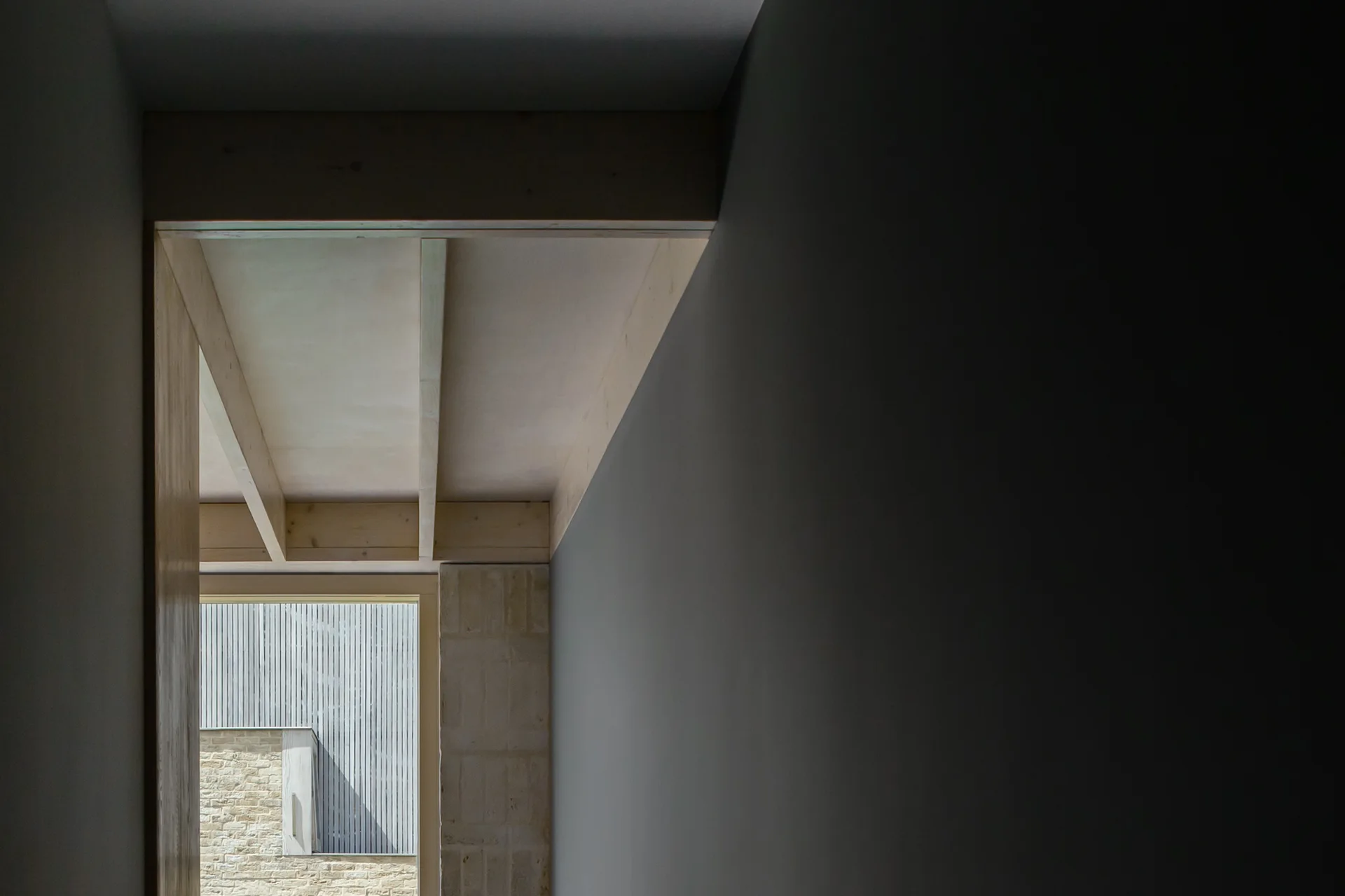 A minimalist corridor with dark grey walls transitions to a bright opening with light wood ceiling joists and a stone wall view.
