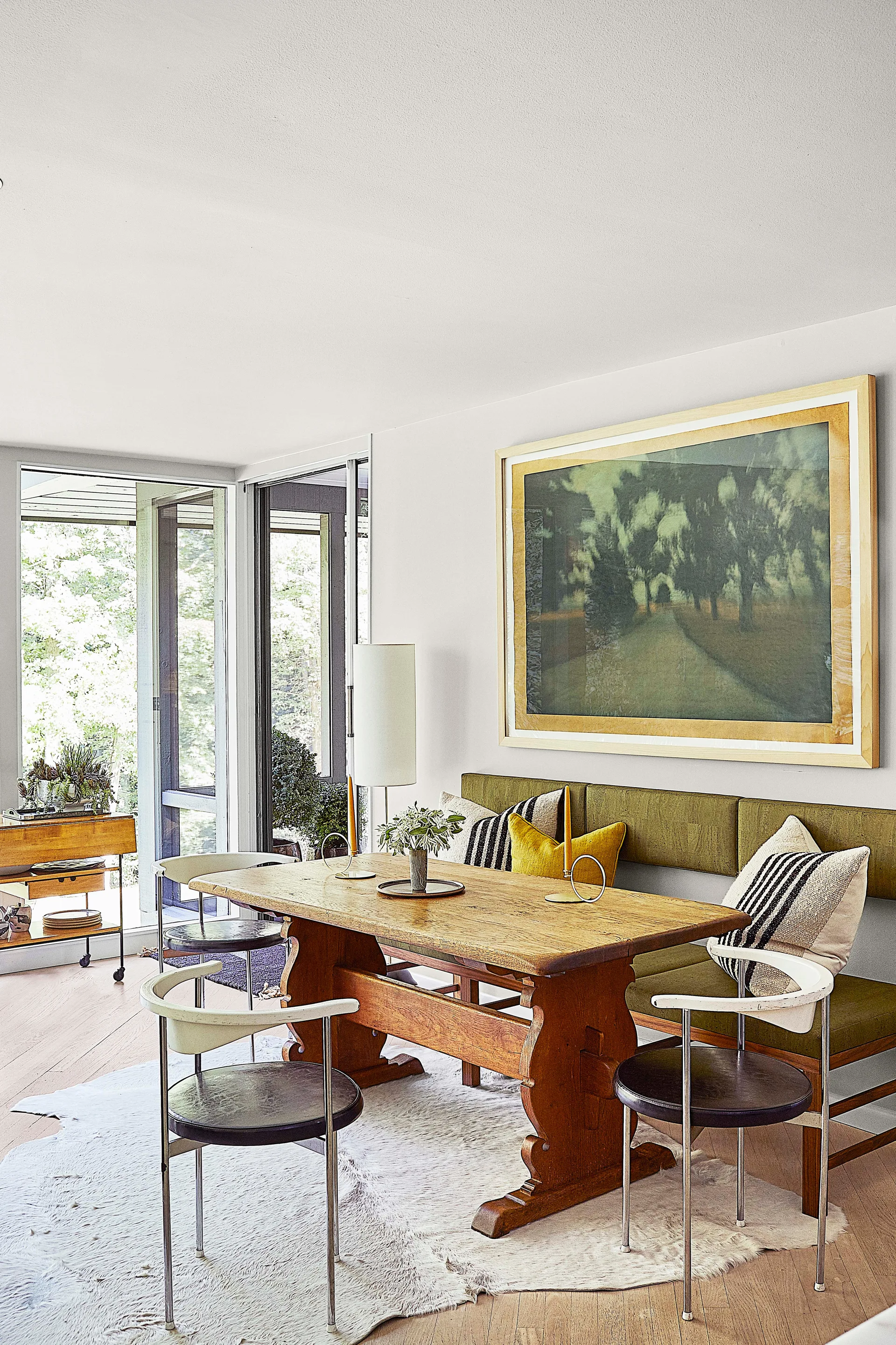 A dining room features a rustic wooden table with metal chairs, an upholstered olive green bench, a cowhide rug, and large framed artwork.