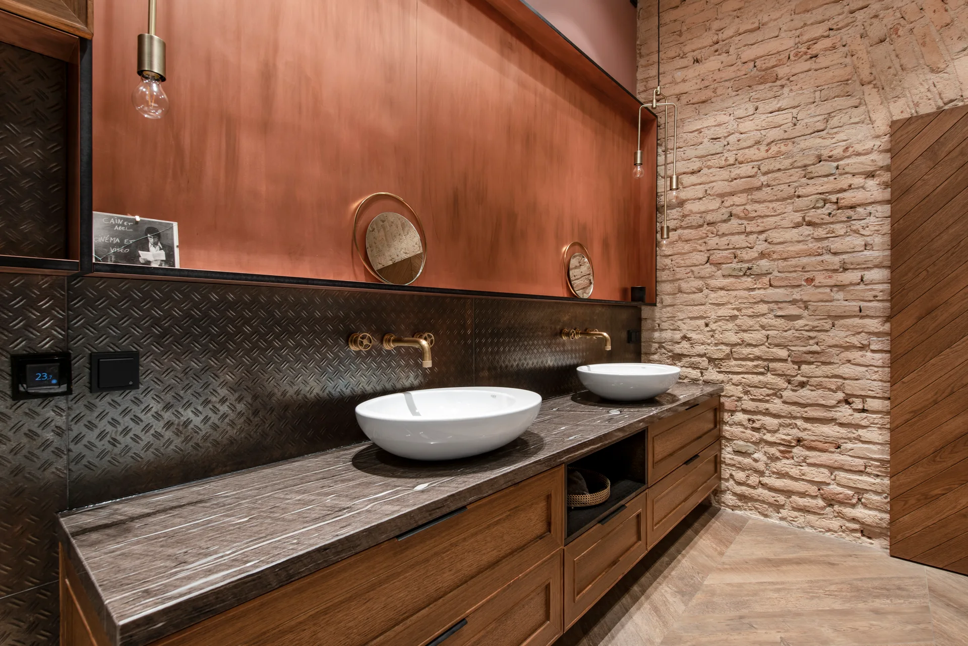 An industrial bathroom vanity features white vessel sinks and brass faucets against a dark textured backsplash, copper wall panel, and exposed brick.
