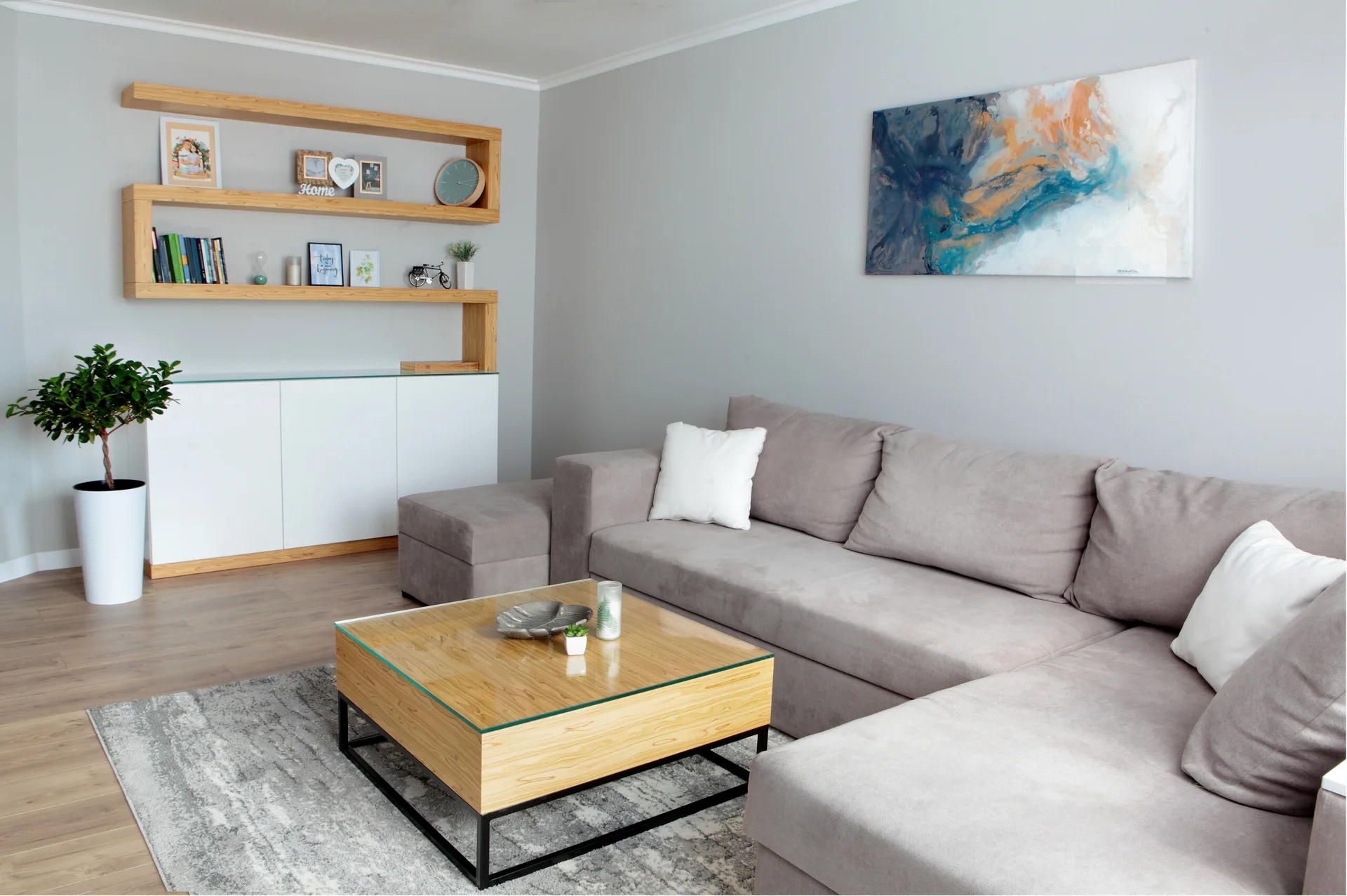 A modern living room features a grey sectional sofa, a glass-top wooden coffee table, and light wood floating shelves displaying books and decor.