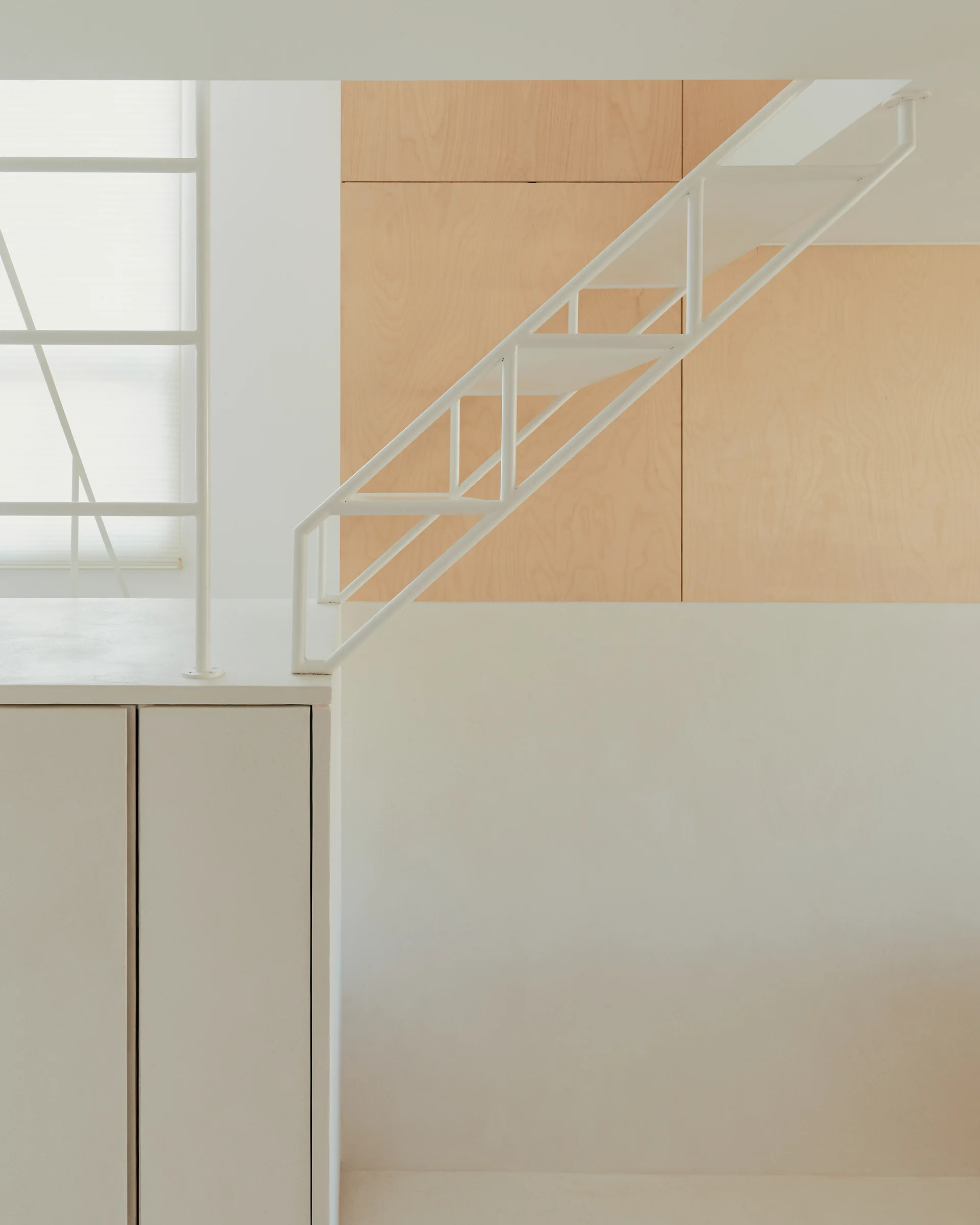 A minimalist white metal staircase stands next to a light wood paneled wall and white cabinetry under a window with a shade.