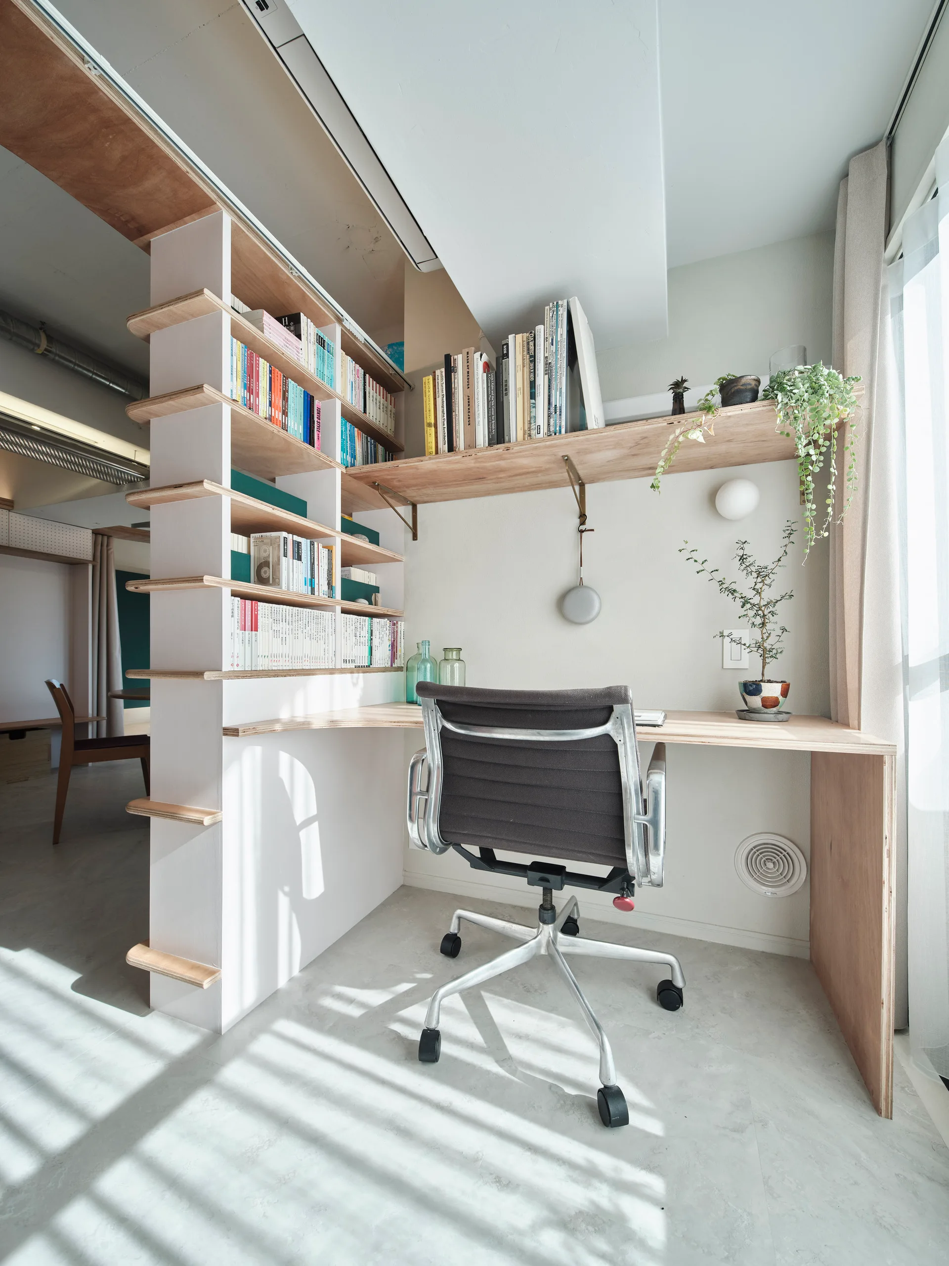 A minimalist home office featuring an integrated plywood desk, built-in shelving unit, black task chair, and sunlit grey floor.