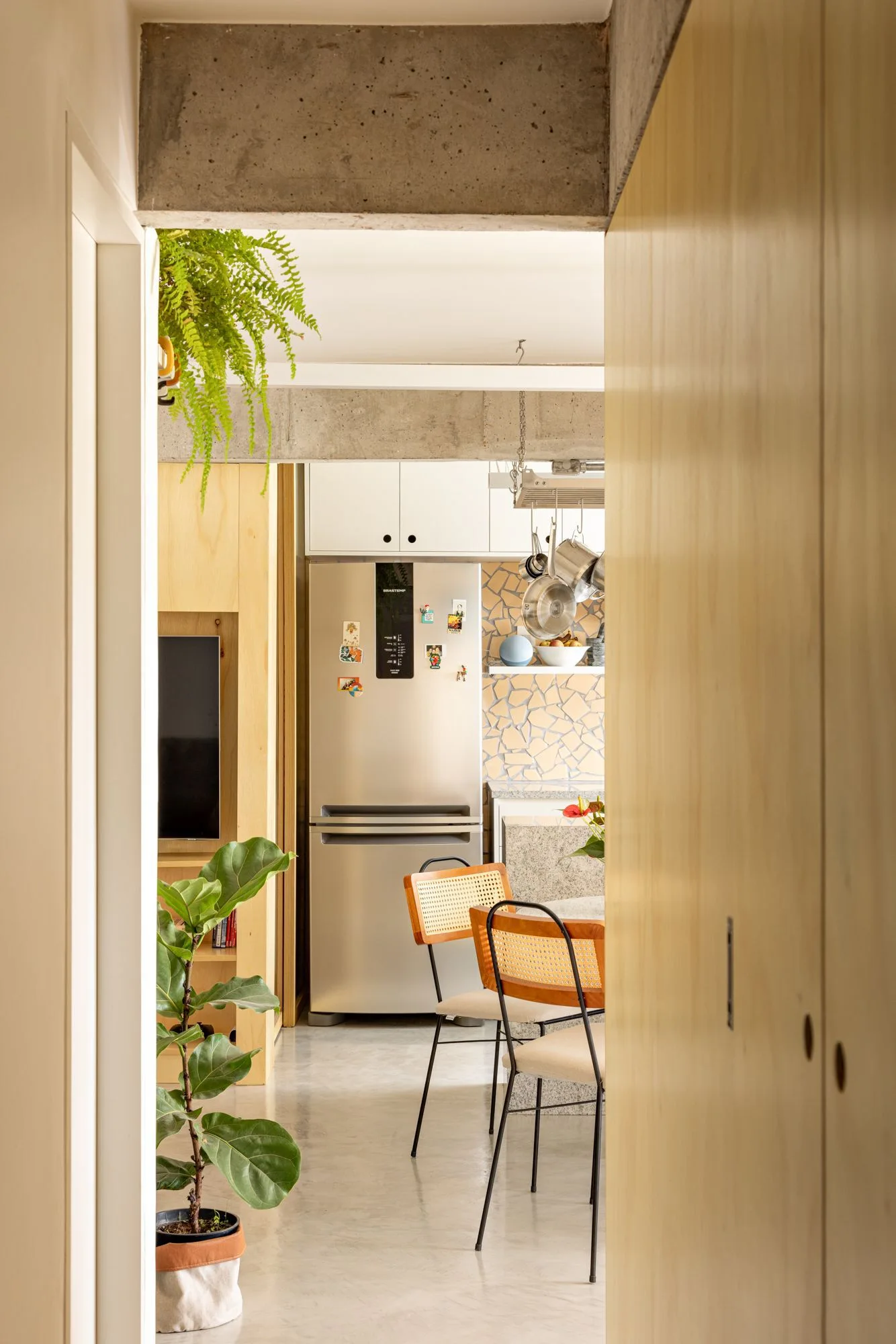 A doorway view into a modern kitchen featuring an exposed concrete beam overhead, cane-back dining chairs, and a stainless steel refrigerator.