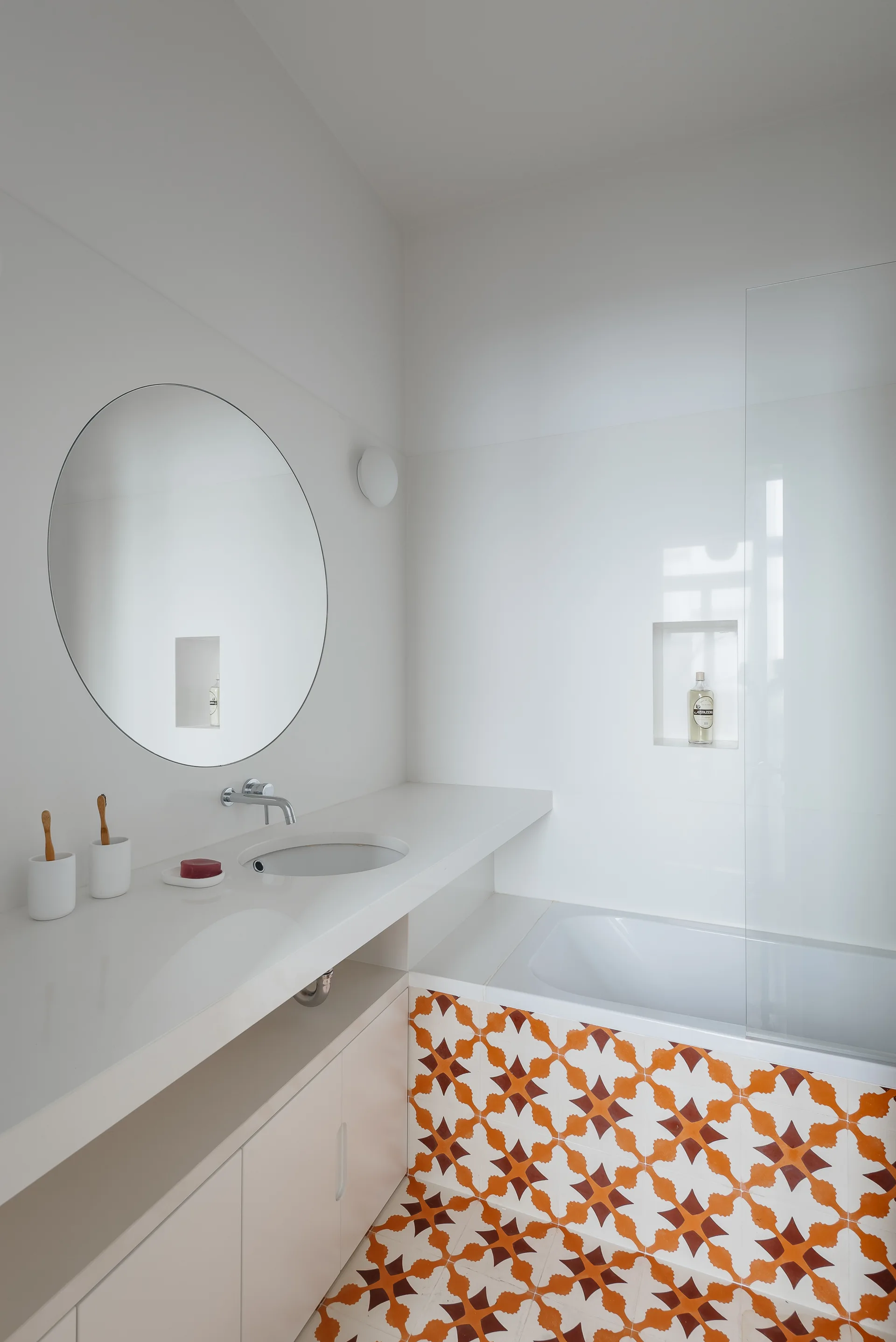 A minimalist white bathroom with a round mirror, an integrated sink vanity, and a bathtub surrounded by orange and white patterned tiles.