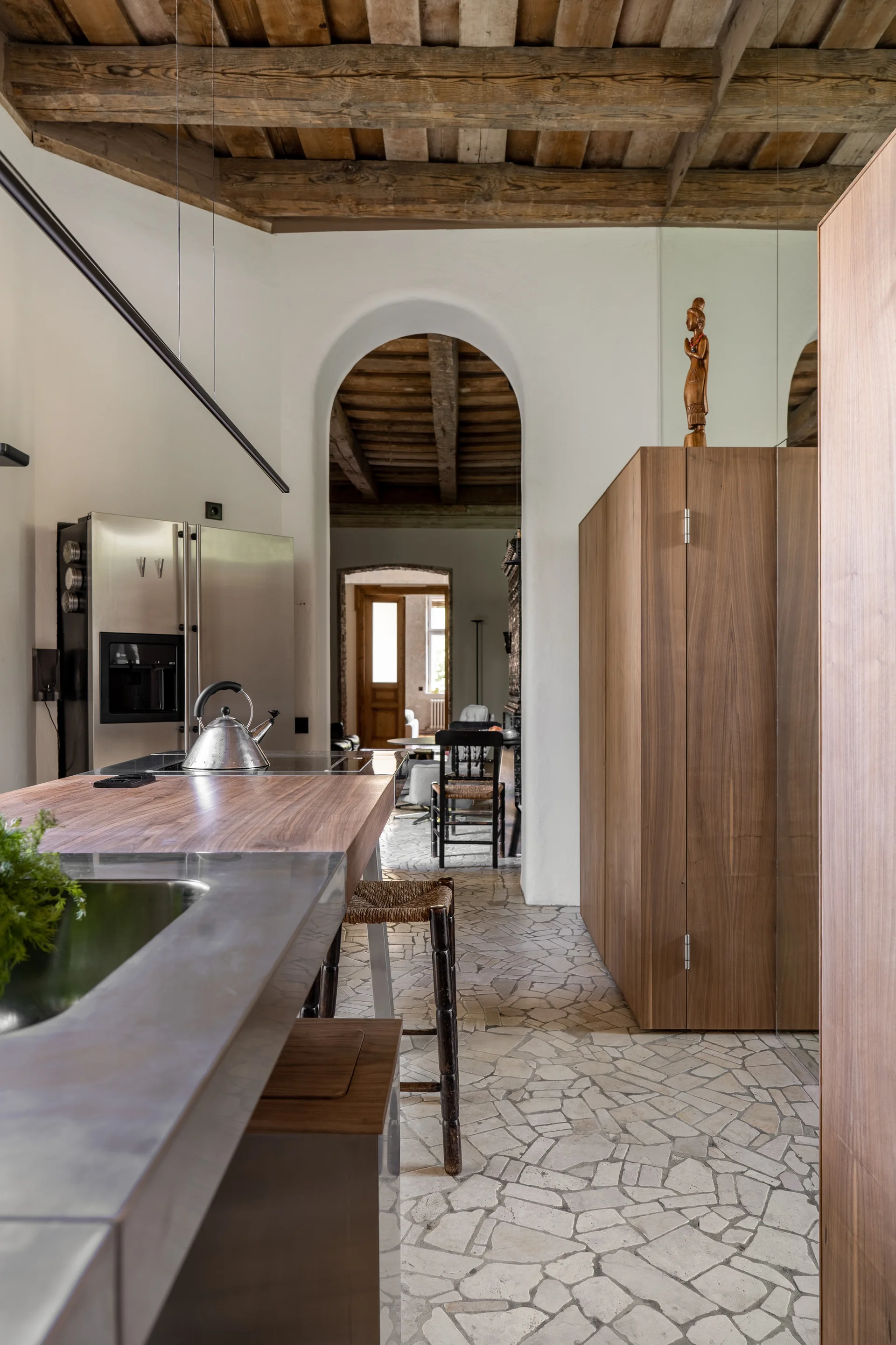 A rustic kitchen features a wood and stainless steel island, irregular flagstone tile floors, and raw wood ceiling beams leading to an arched doorway.