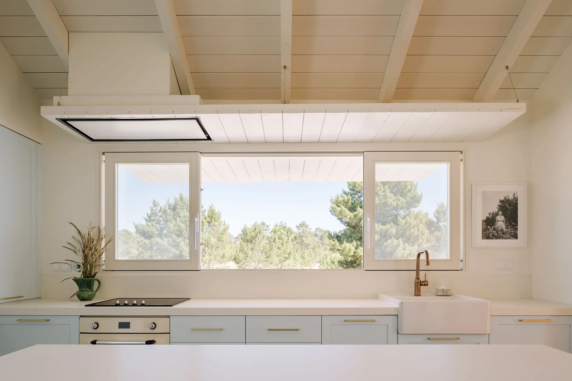 A bright kitchen features pastel blue lower cabinets, a farmhouse sink with a brass faucet, and a large window showcasing a view of pine trees.