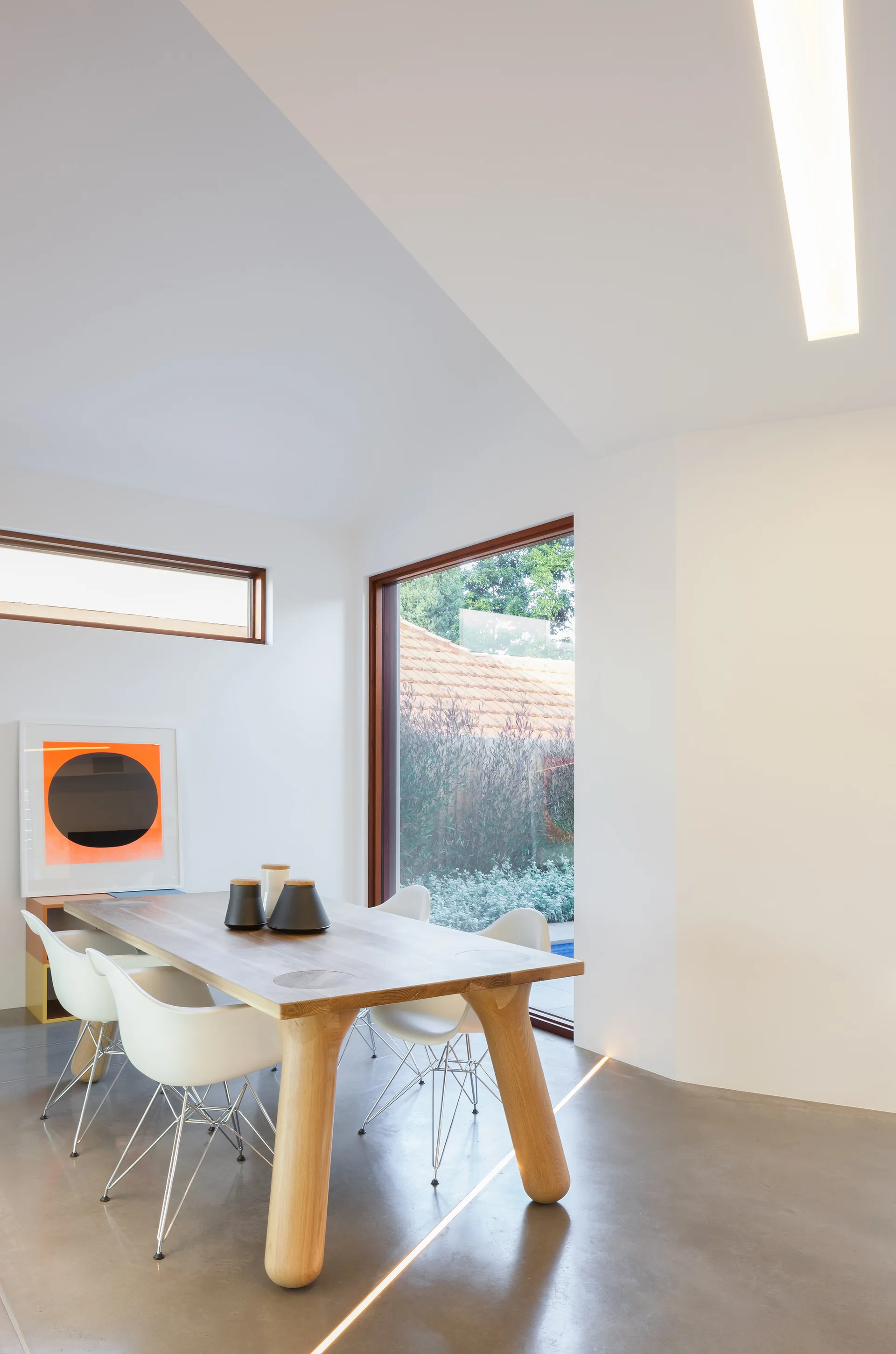 A modern dining room featuring a light wood table, white shell chairs, polished concrete floors with an integrated light strip, and large windows.
