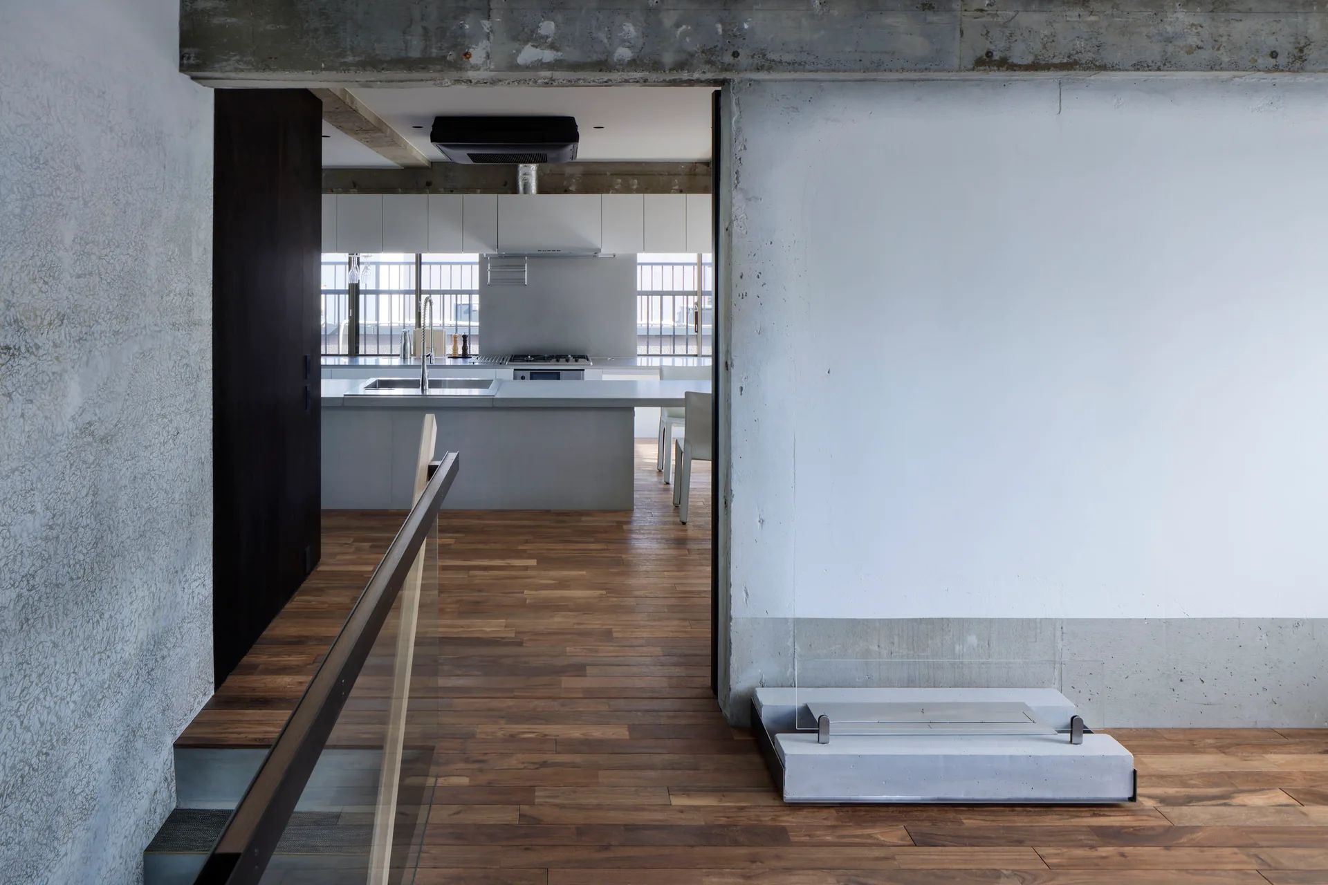 A minimalist kitchen with a gray island is seen through an opening next to a low concrete fireplace on rich wood floors.