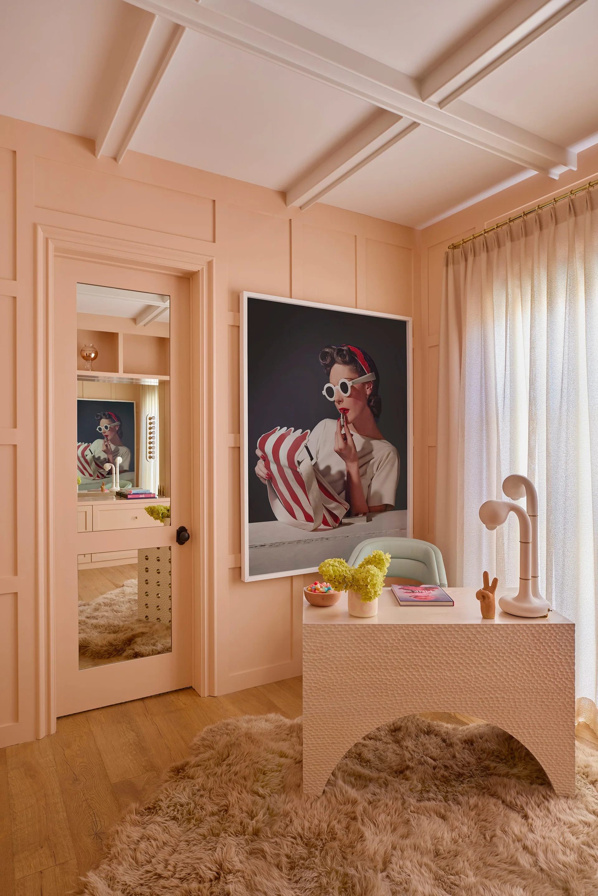 A monochromatic pink home office featuring a textured arched desk, a shaggy tan rug, and a large framed pop art portrait on a paneled wall.