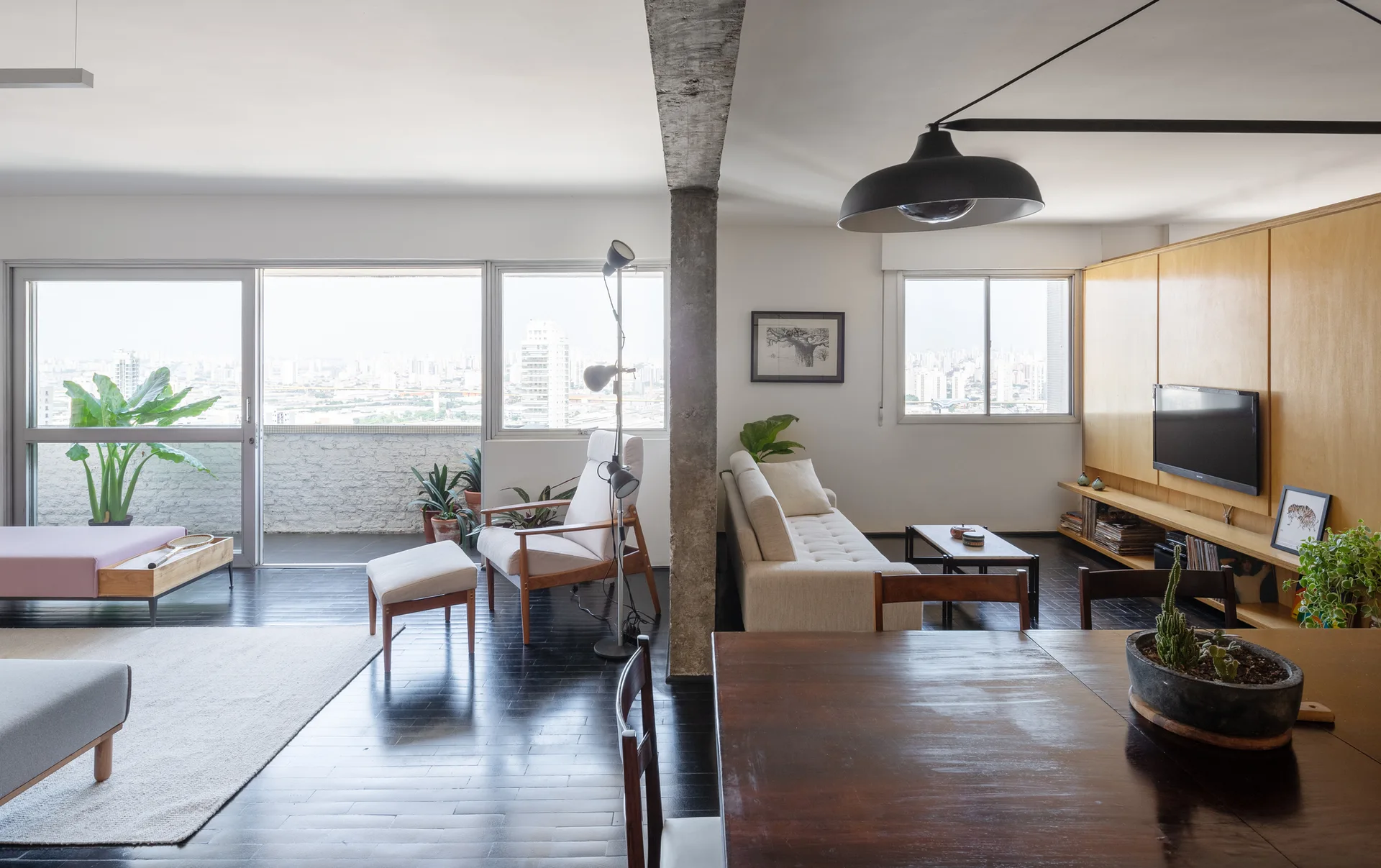 An open-plan apartment living and dining area features dark wood floors, a central exposed concrete column, and a large wooden wall cabinet.