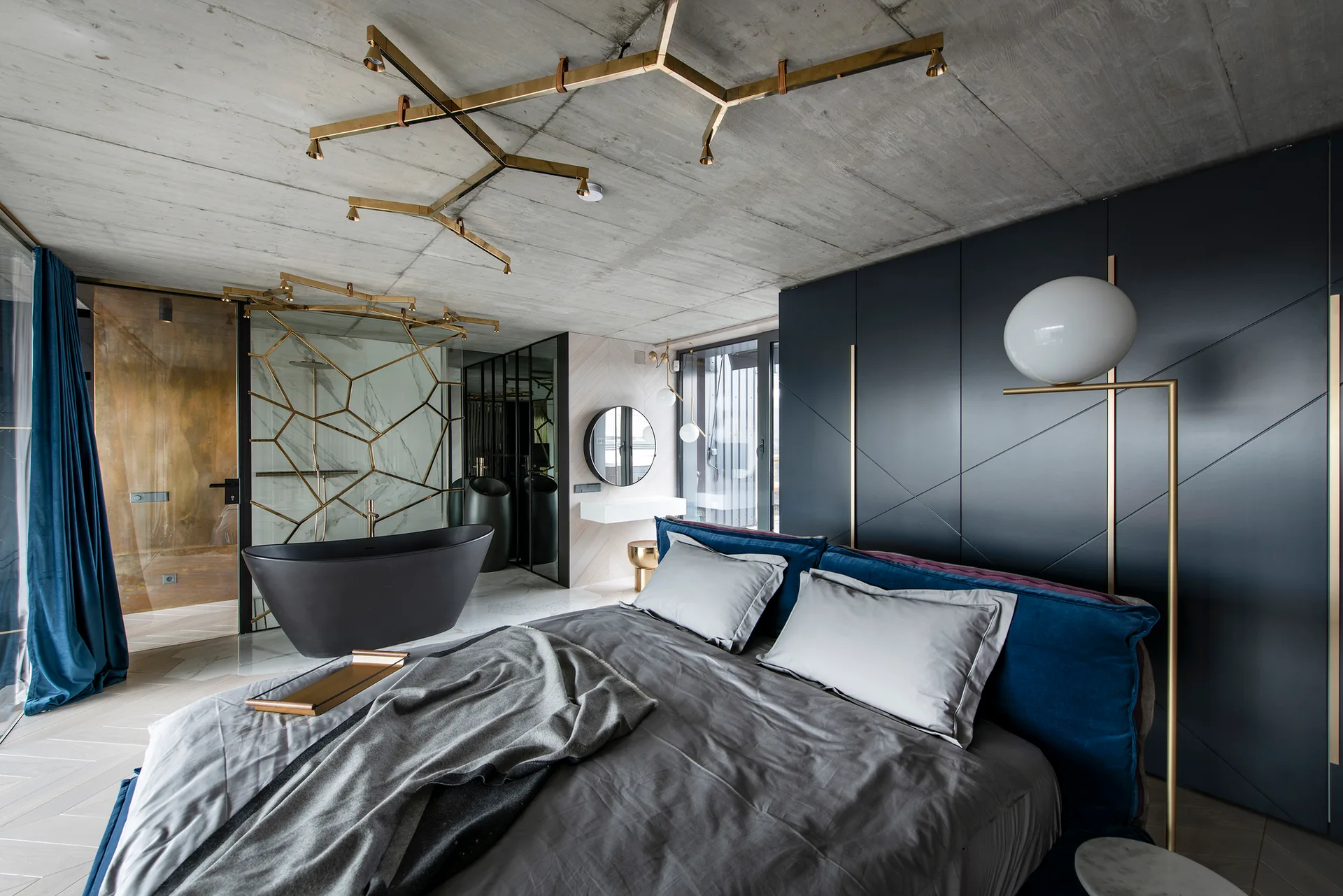 Industrial bedroom featuring a blue bed, raw concrete ceiling, dark grey paneling, and an open ensuite with a black freestanding bathtub.