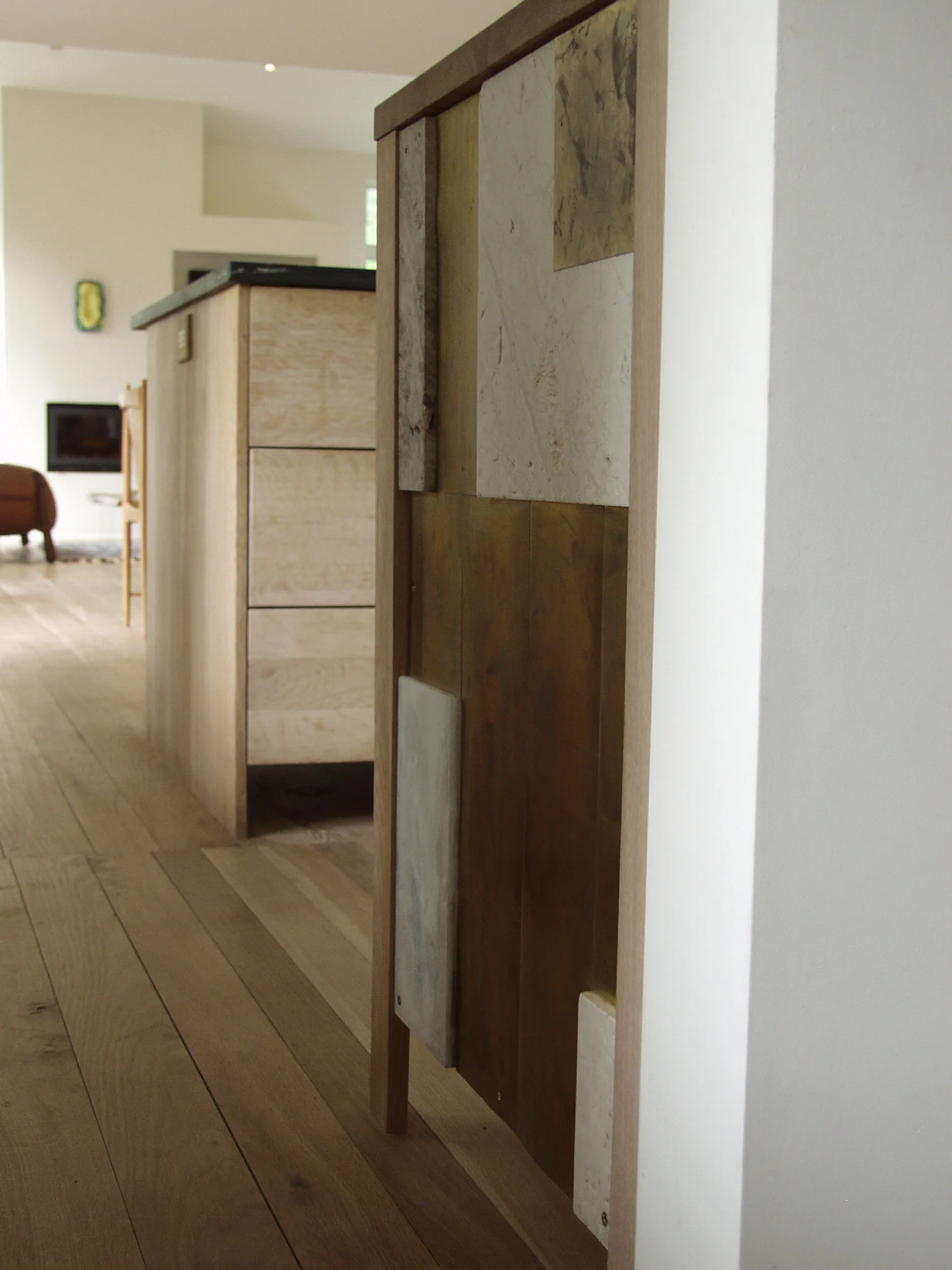 A close-up of a modern cabinet panel made from patchwork wood and stone tiles, with a light wood kitchen island and hardwood floors in the background.