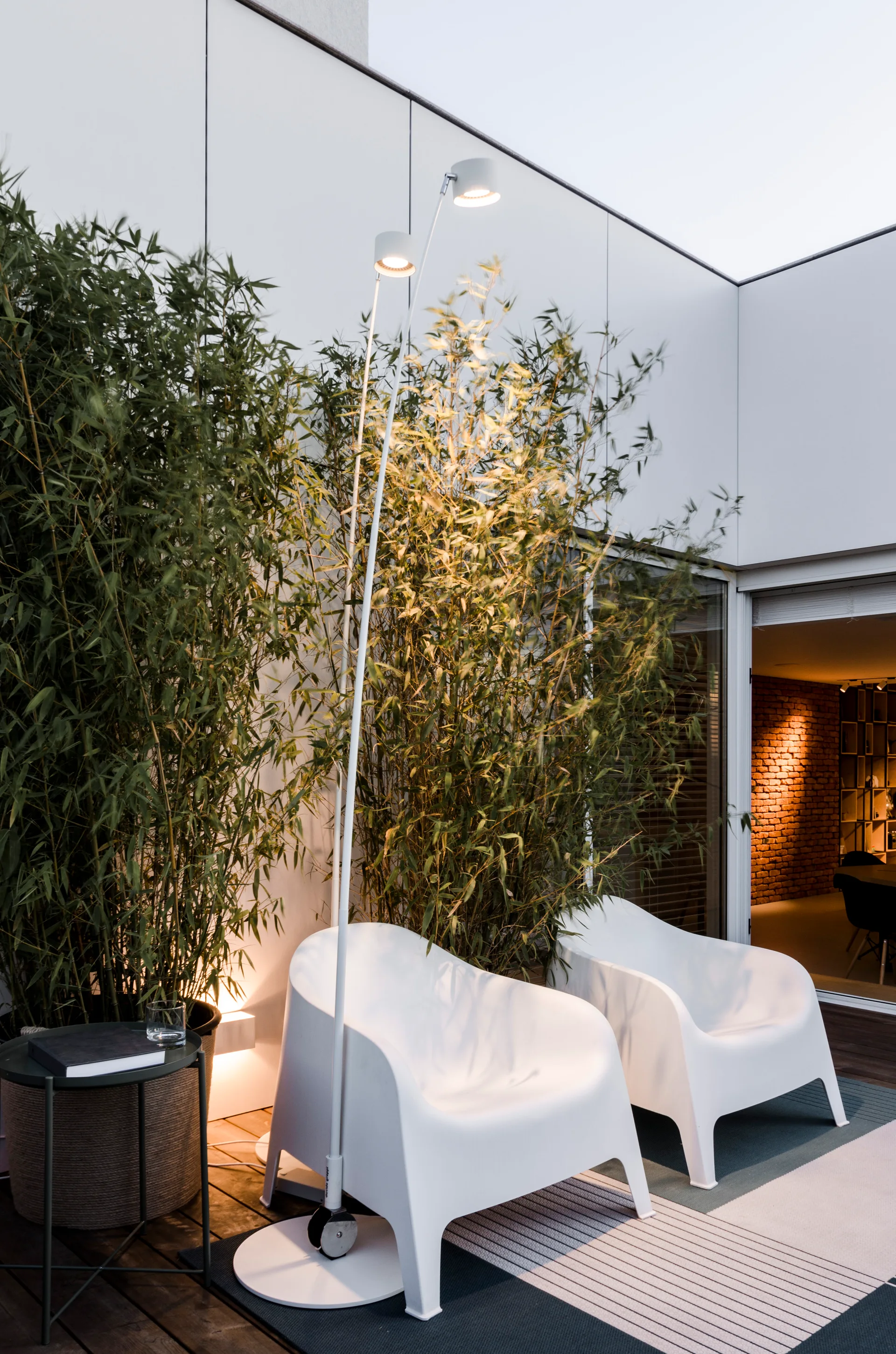 Two white plastic lounge chairs and a tall floor lamp sit on a dark wood patio deck next to tall green bamboo plants.